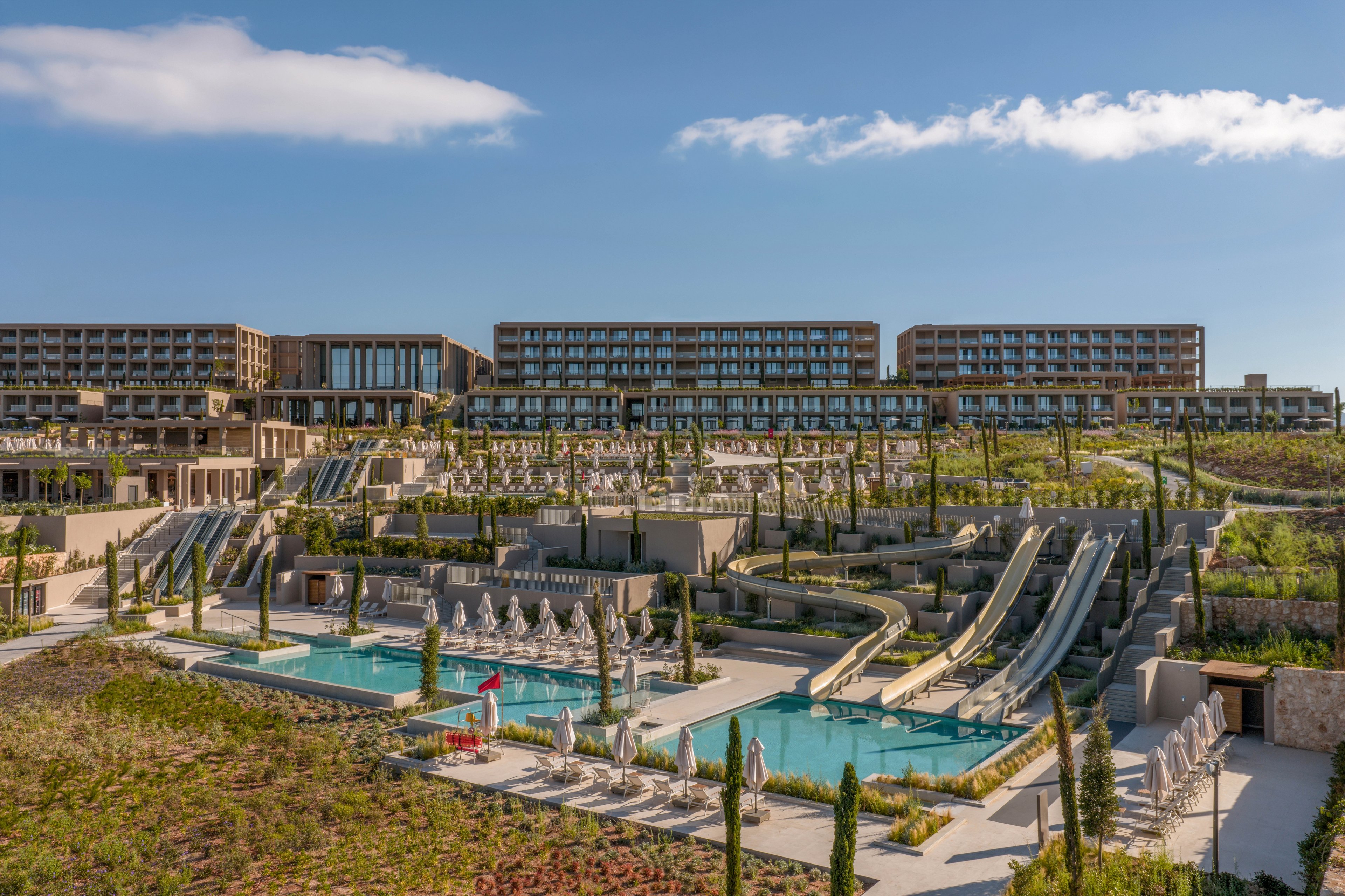 A large contemporary resort complex is shown with multiple terraced buildings overlooking landscaped grounds. In the foreground, several outdoor swimming pools feature rows of sun loungers and umbrellas. Multiple parallel waterslides descend toward the pools, suggesting a water park style recreation area. The scene is captured under a clear blue sky with scattered clouds, creating a bright and inviting vacation atmosphere.