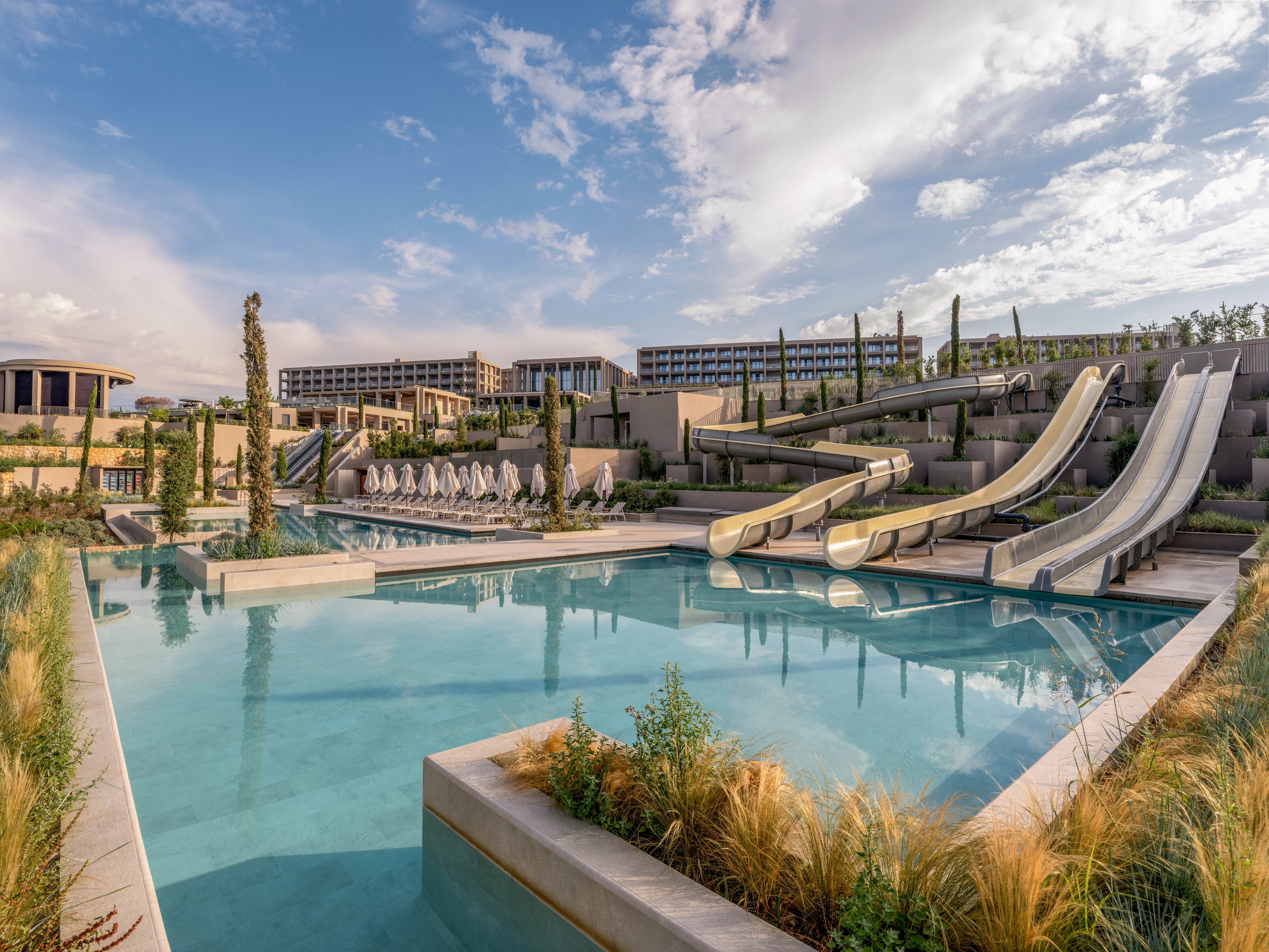 A large outdoor pool complex features multiple tall water slides leading into clear blue water. In the background, a modern resort building and rows of white sun loungers with umbrellas line the poolside. Decorative grasses and landscaped greenery frame the foreground, adding a natural touch to the contemporary setting. The sky is partly cloudy, creating a bright and inviting atmosphere for leisure and vacation.