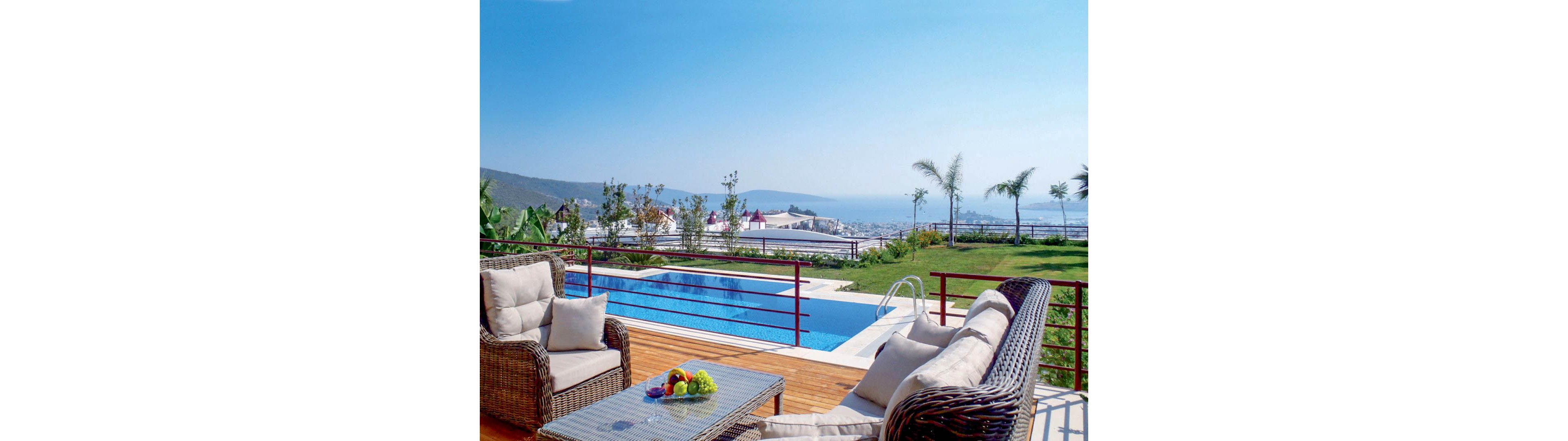 An elegant outdoor terrace features wicker lounge furniture arranged around a glass-top table with a bowl of fresh fruit. Beyond the seating area, a rectangular swimming pool sits on a wooden deck, bordered by a metal railing. The scene overlooks a coastal town and calm blue sea under a clear sky, with distant hills and scattered buildings visible. Palm trees and manicured grass add a resort-like atmosphere to the tranquil setting.