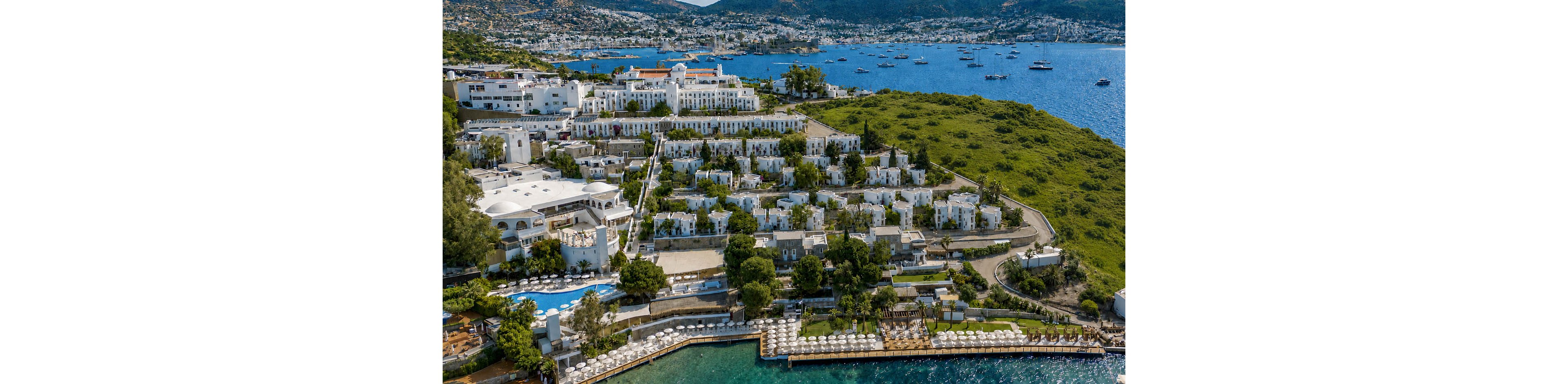 An expansive coastal resort complex is captured from above on a sunny day. White buildings and villas are arranged along a green hillside that extends into a blue bay. Multiple swimming pools, sun loungers, and terraces line the waterfront. In the background, boats and yachts are anchored across the calm sea with a distant shoreline and hills.