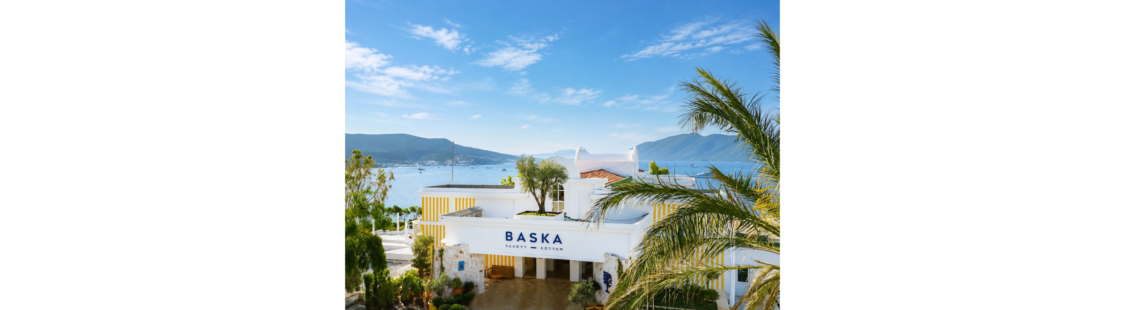 A coastal resort building labeled "BASKA" stands in the foreground, framed by lush palm trees and greenery. Behind the property, a calm blue sea stretches out toward distant mountains under a clear sky. Yellow and white striped architectural accents add a bright, summery touch to the scene. Boats are visible on the water, enhancing the relaxed vacation atmosphere.