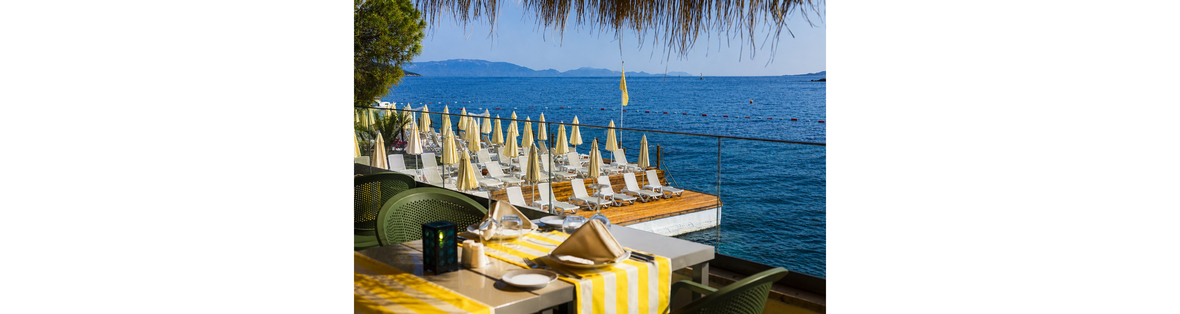 A seaside resort terrace dining area overlooks rows of empty sun loungers and umbrellas by the water. The foreground shows a neatly set table with striped yellow and white table runners, napkins, and glassware. Beyond the glass railing, a wooden deck with loungers leads to the calm blue sea marked by a line of buoys. Distant hills and a clear sky create a bright, relaxing holiday atmosphere.