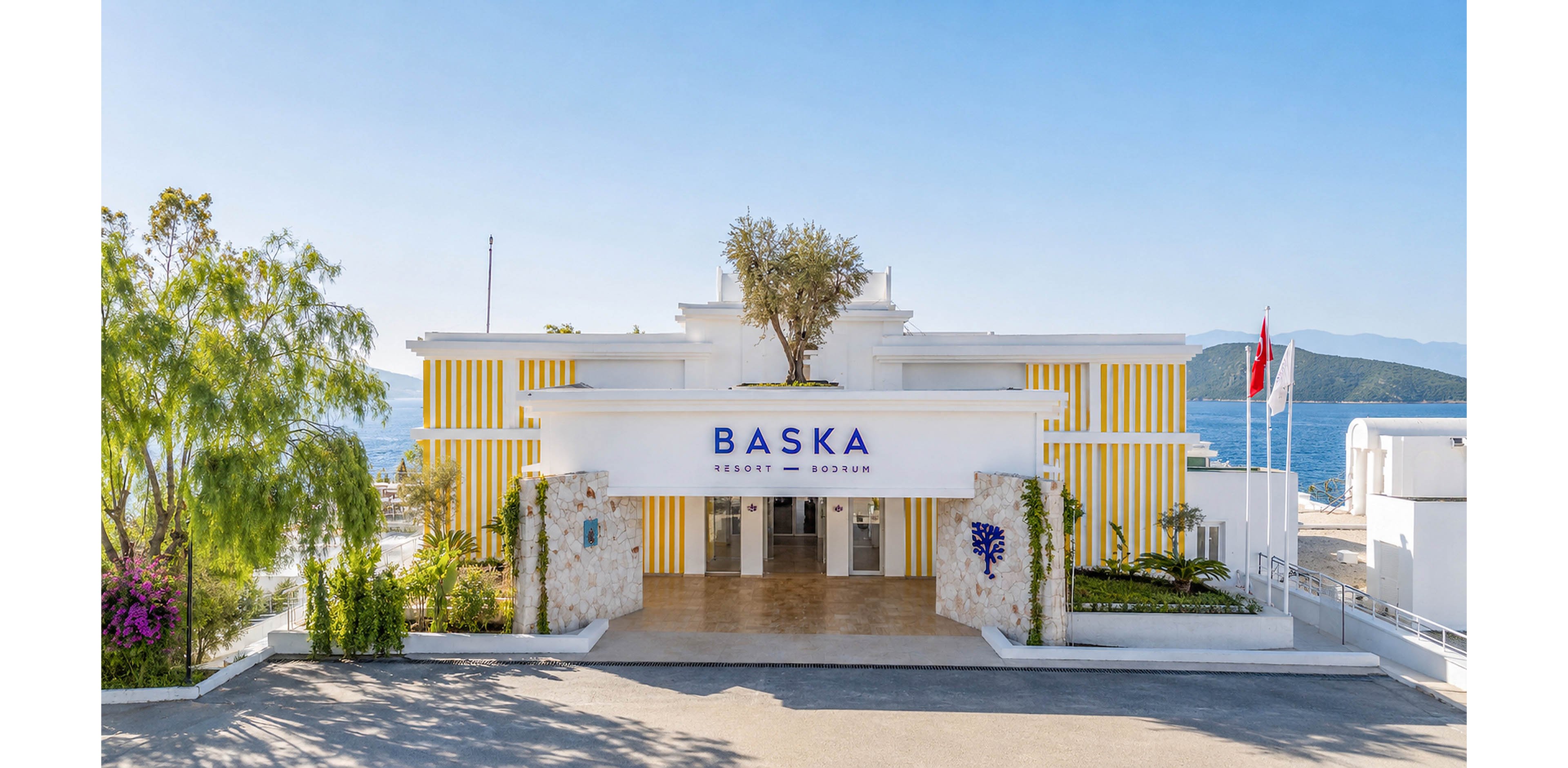 A modern seaside resort building labeled BASKA stands prominently with the ocean visible in the background. The structure features clean white walls, yellow vertical accents, and a central tree above the main entrance. Decorative stone planters with purple flowers and lush greenery frame the entryway. A clear blue sky and distant hills emphasize the coastal setting and relaxed vacation atmosphere.