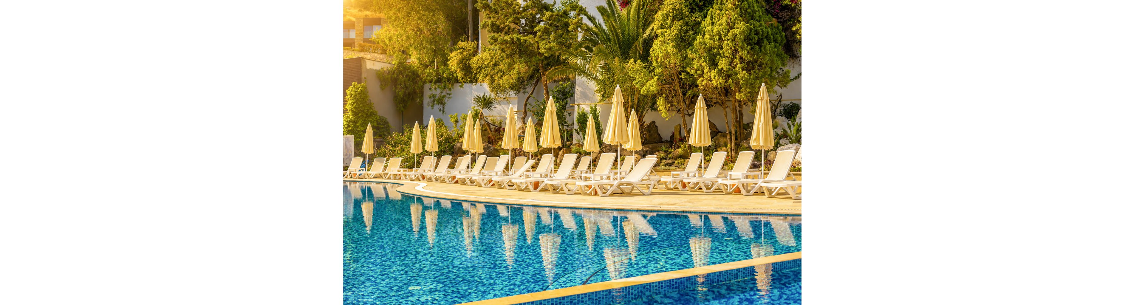 A row of white sun loungers with beige umbrellas lines the edge of a clear blue swimming pool. The scene is bathed in warm golden sunlight, suggesting early morning or late afternoon at a resort. Lush green trees and palm trees form a backdrop, creating a tropical and tranquil atmosphere. The absence of people emphasizes relaxation and calm in this vacation setting.