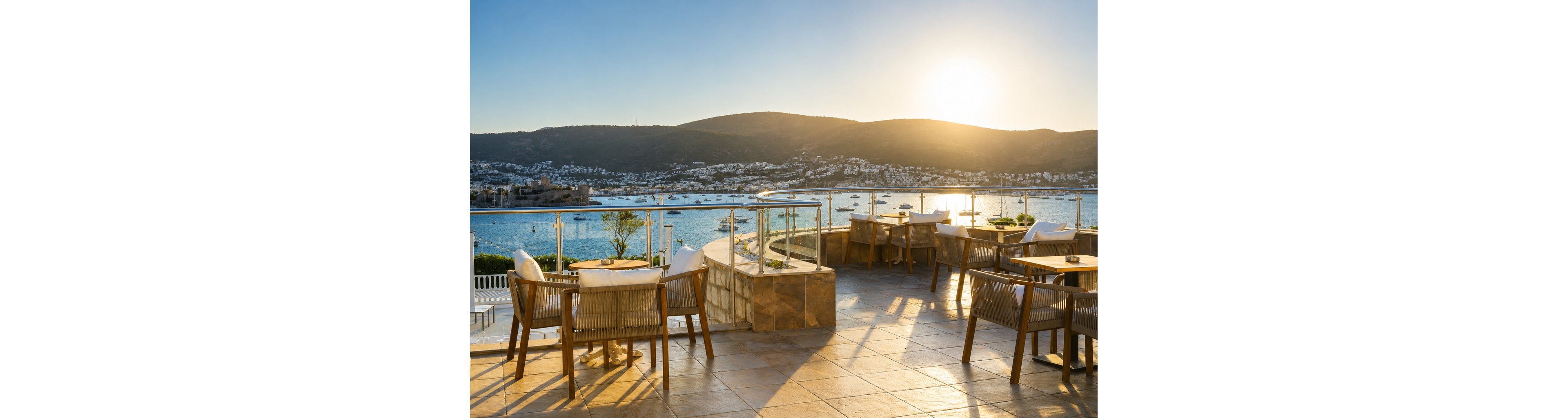 An open-air terrace restaurant overlooks a calm coastal bay at sunset. Wooden tables and chairs are arranged along a stone-tiled patio with glass railings. The sun is low over distant hills, casting warm golden light across the water. White buildings and numerous small boats dot the shoreline, creating a tranquil holiday atmosphere.