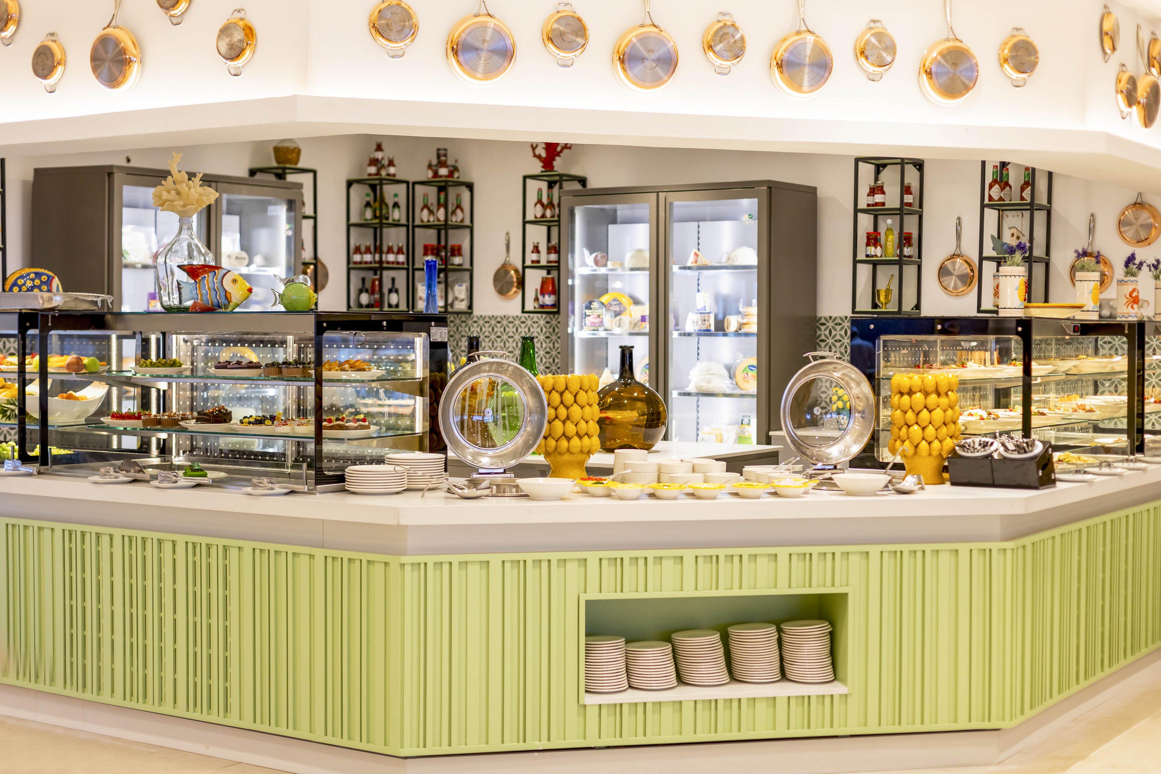 A brightly lit dessert buffet counter is arranged with various pastries and sweets. Glass display cases and refrigerated cabinets showcase cakes, tarts, and other treats. Large jars of lemons and decorative plates on the wall add a fresh, colorful accent. The counter front is painted a soft green, with neatly stacked plates ready for guests.
