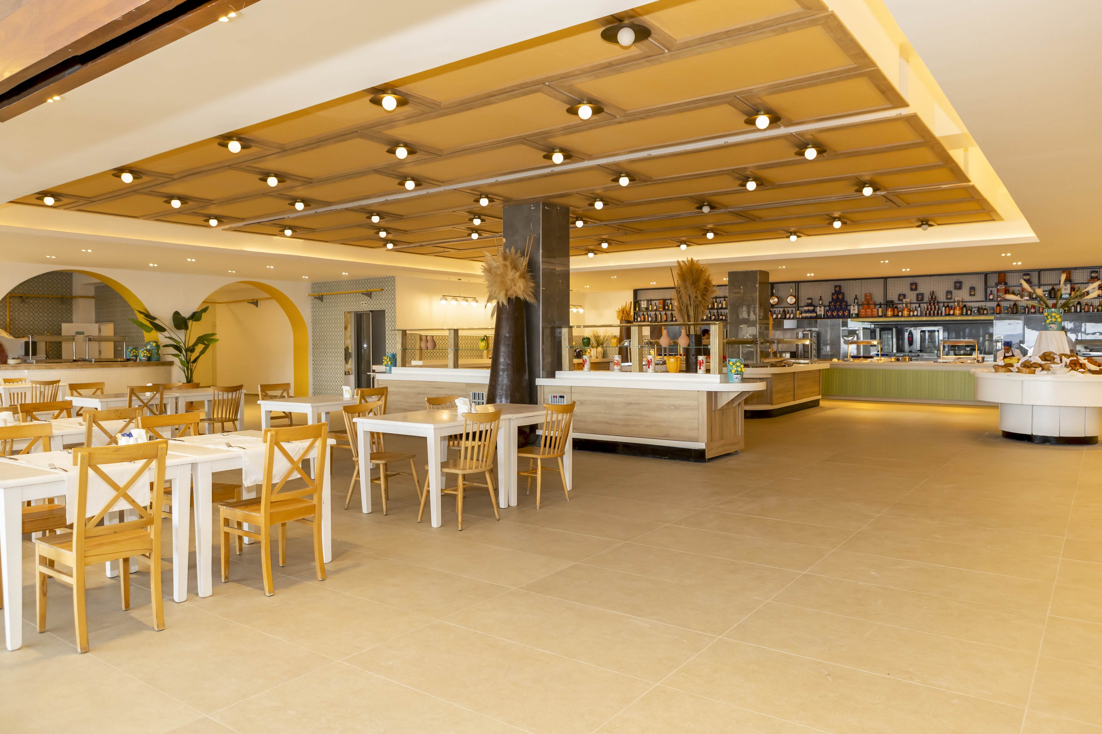 A spacious, modern cafeteria interior features light wooden tables and chairs arranged neatly across a tiled floor. The ceiling is lined with warm recessed lights, creating an inviting atmosphere. In the background, a long self-service buffet counter and beverage area are visible, with shelves of bottles and decor. Neutral beige and yellow tones dominate the space, giving it a clean and contemporary look.