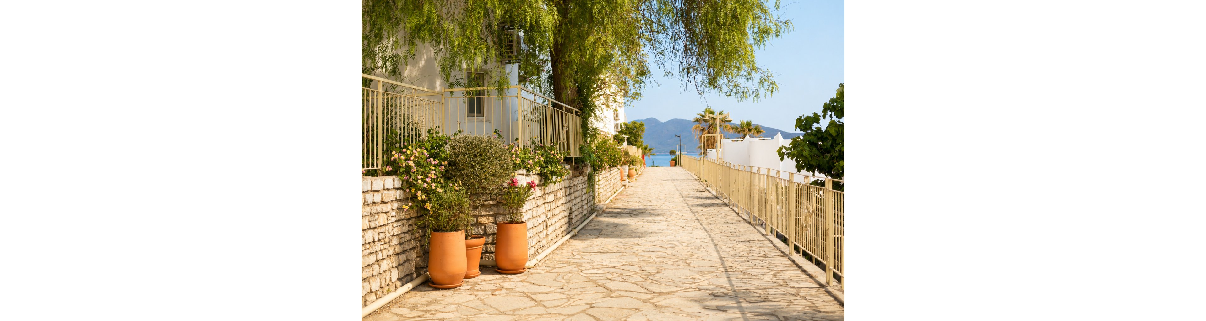 A sunlit stone walkway runs between a railing and a low stone wall lined with terracotta flower pots. Lush green plants and blooming flowers add color and texture along the path. In the distance, blue water and hazy mountains suggest a coastal setting. The overall scene feels warm, quiet, and inviting, evoking a relaxed Mediterranean atmosphere.