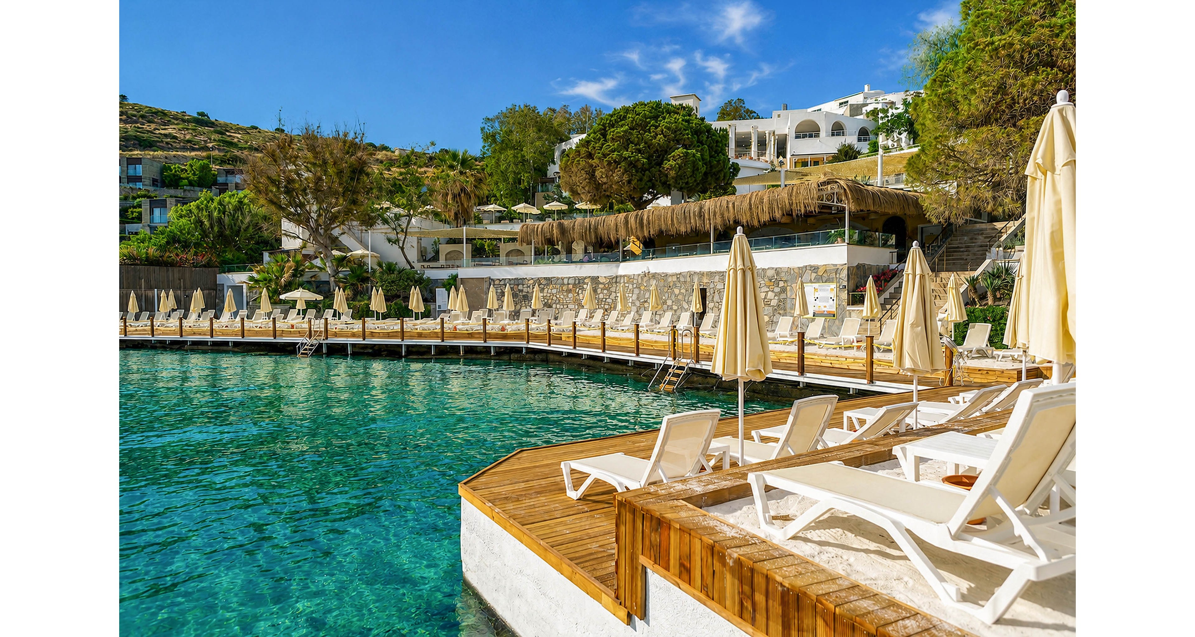 A tranquil seaside resort features rows of empty sun loungers and closed umbrellas on a wooden deck. Clear turquoise water gently meets the shoreline, reflecting the bright blue sky above. White buildings and lush greenery rise on the hillside in the background, creating a Mediterranean-style setting. The scene feels calm and inviting, ideal for a relaxing beach holiday.