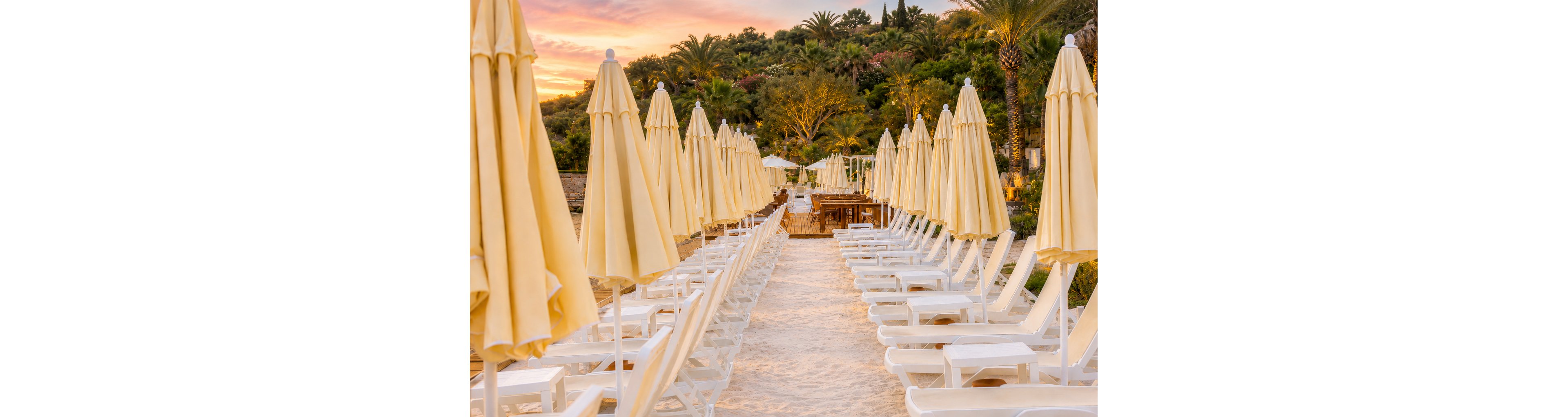 Rows of white sun loungers with closed beige umbrellas line a sandy beach pathway. The scene is empty of people, creating a calm and orderly seaside setting. Lush green palm trees and vegetation frame the background under a colorful sunset sky. Warm golden light enhances the tranquil vacation atmosphere.