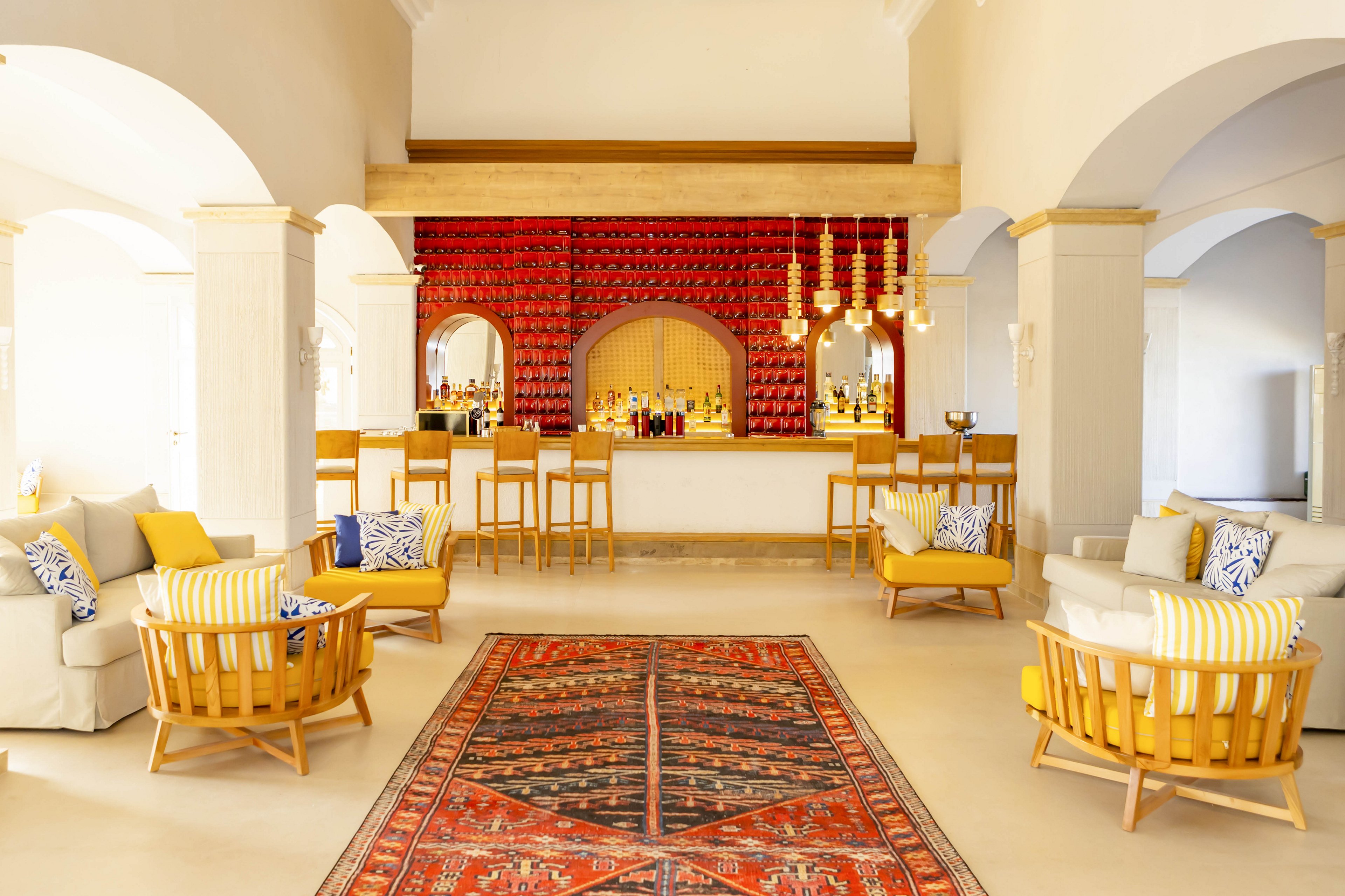 A spacious hotel lounge features a central bar framed by arches and warm neutral walls. Wooden chairs with yellow cushions and patterned pillows surround low tables, creating a relaxed seating area. A red patterned rug runs down the center of the room toward the bar, adding color and texture. Pendant lights hang above the bar, enhancing the inviting, contemporary atmosphere.