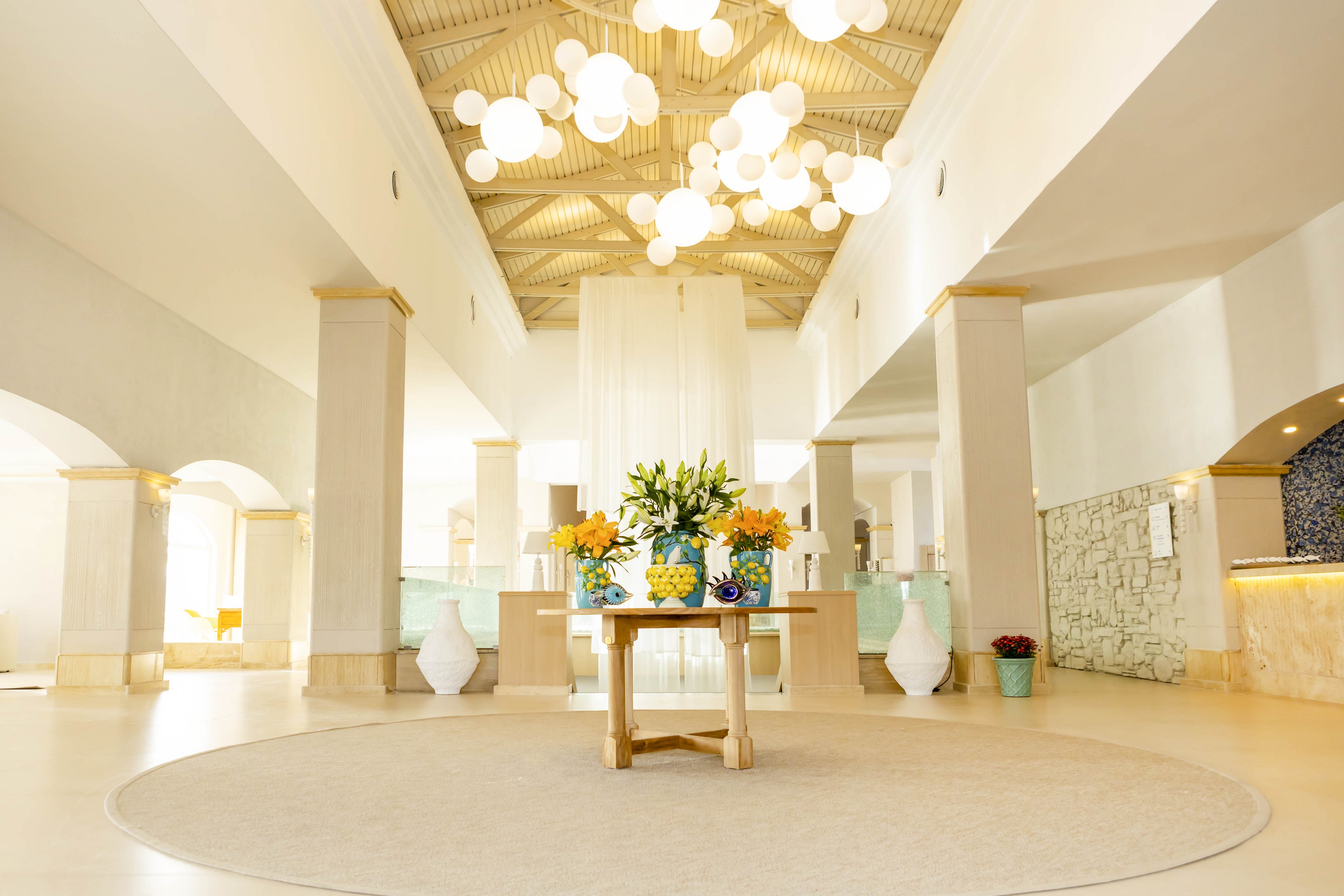 A spacious, modern hotel lobby features a wooden table with vibrant flower arrangements at its center. The room is illuminated by numerous round pendant lights hanging from a high, vaulted ceiling. Soft neutral tones dominate the walls and floor, complemented by light green glass accents at the reception area. Large decorative vases and subtle architectural details create an elegant, welcoming atmosphere.