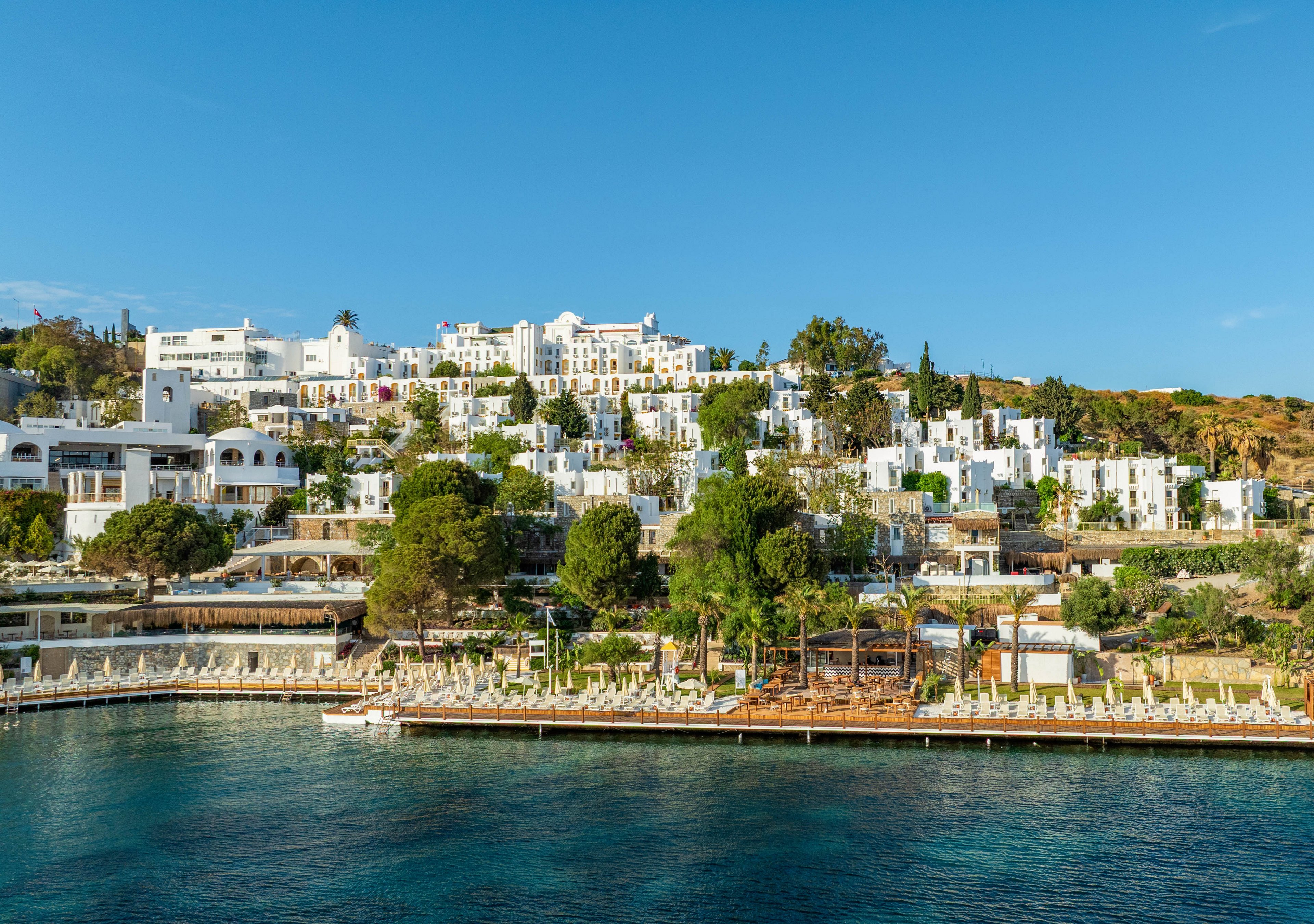 A terraced cluster of white buildings lines a coastal hillside above a calm blue sea. The scene appears to show a resort or residential complex with multiple levels of structures and greenery. Sun loungers and umbrellas are arranged along the waterfront, suggesting a beach or pool area. Clear skies and bright sunlight create a warm, inviting atmosphere.