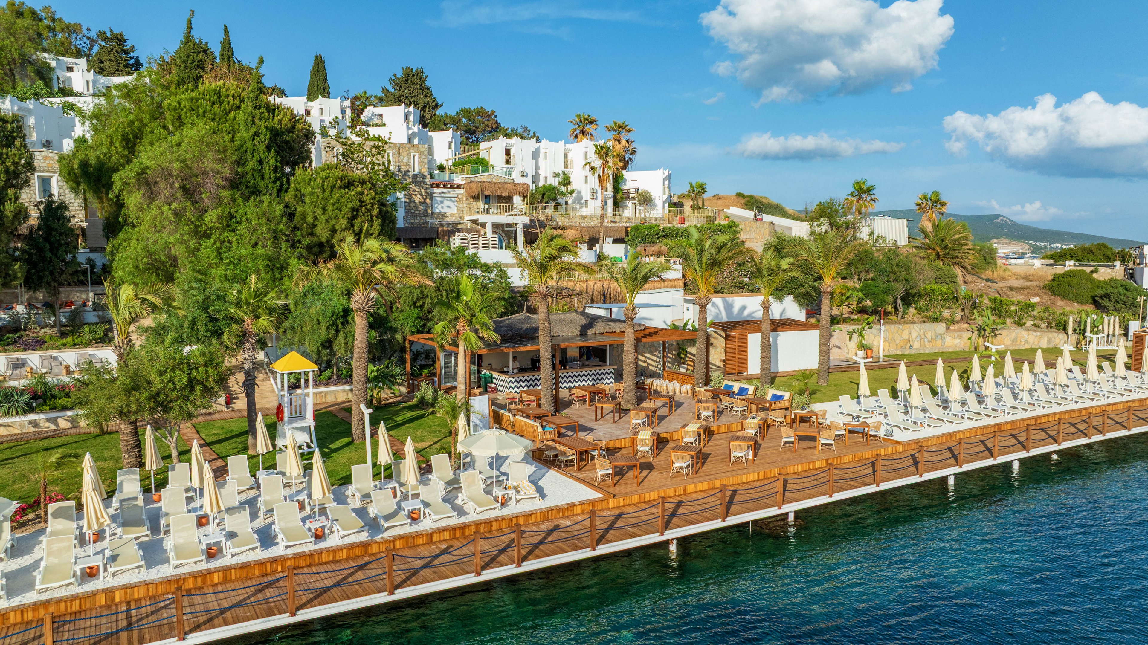 A coastal resort features a long wooden deck lined with neatly arranged sun loungers and umbrellas beside calm blue water. Palm trees and lush greenery frame the outdoor seating areas, creating a tropical atmosphere. White hillside buildings rise in the background, suggesting a Mediterranean or coastal village setting. The clear sky with scattered clouds enhances the bright, relaxed vacation mood.