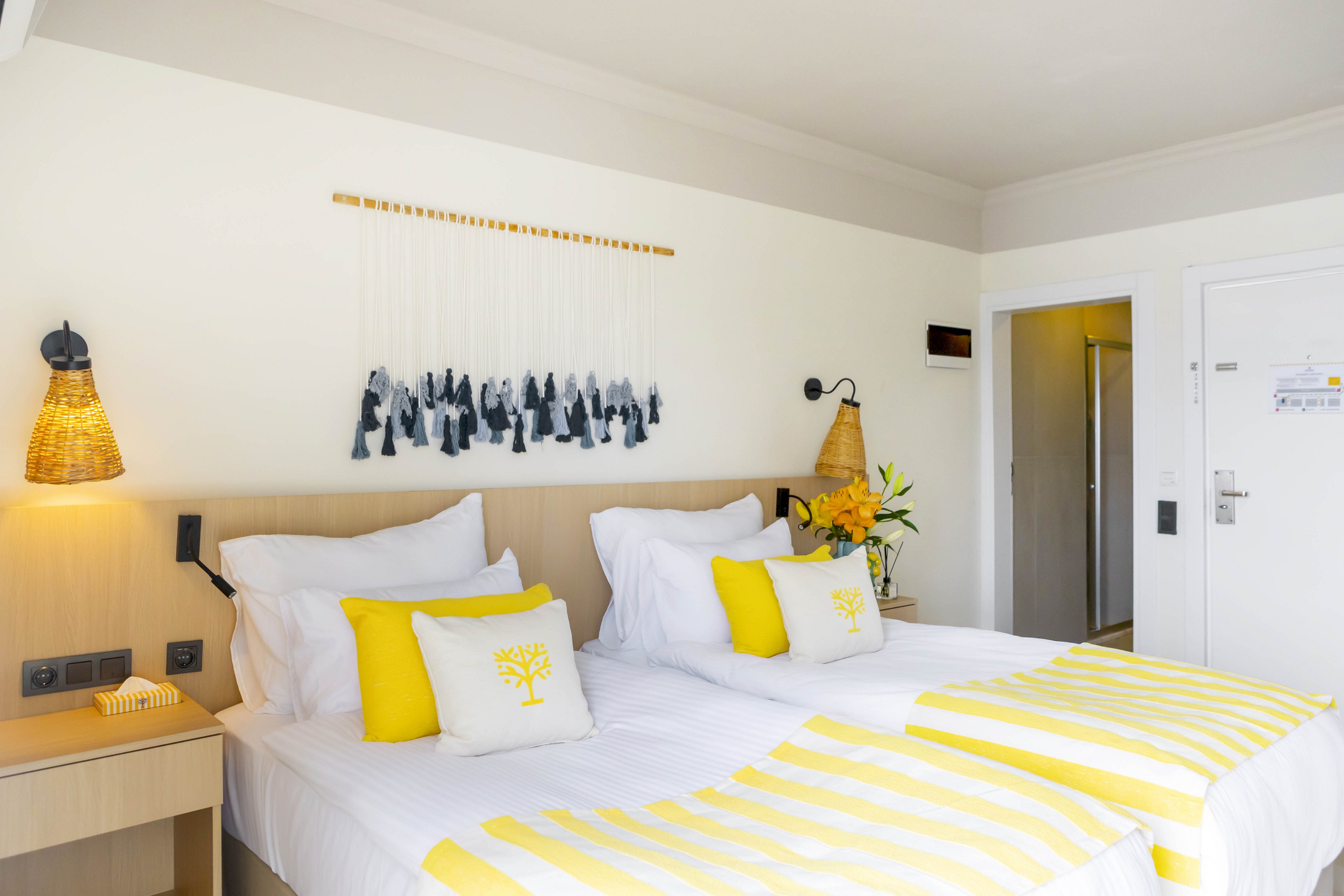 A bright, modern hotel room features two neatly made twin beds with white linens and yellow striped throws. Coordinated yellow accent pillows and a small bouquet of flowers add warmth and color to the neutral space. Woven bedside lamps, a wooden headboard, and minimalist wall art contribute to a clean, contemporary style. A bedside table, wall switches, and an open closet area are visible, emphasizing the room’s functional layout.