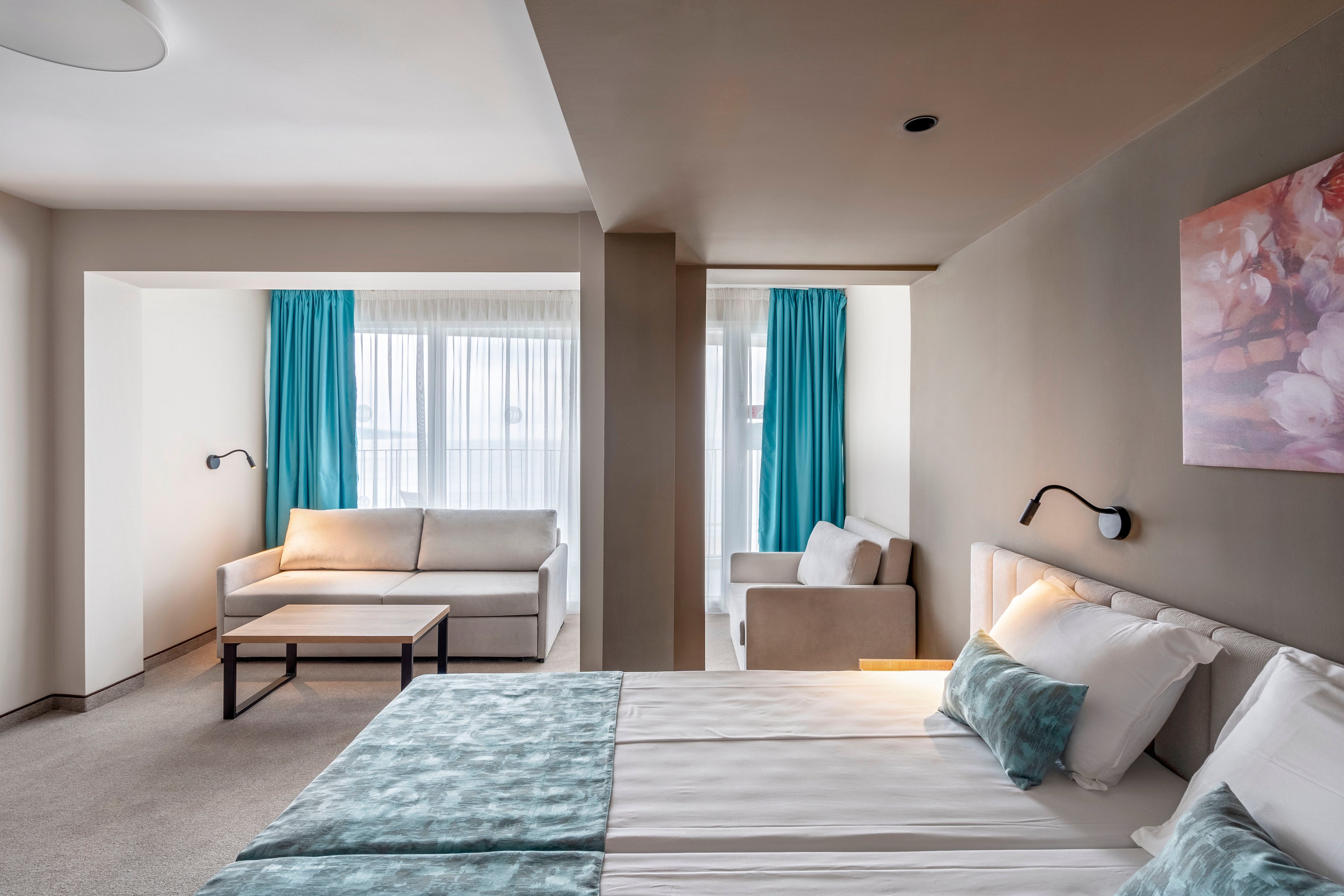 Bright contemporary hotel room featuring a double bed with light blue accents. A cozy sofa seating area is positioned near large windows with sheer curtains and turquoise drapes. Neutral walls and carpet create a calm, minimalist atmosphere, complemented by a floral artwork above the bed. Wall-mounted reading lights and a simple coffee table add functional comfort to the space.