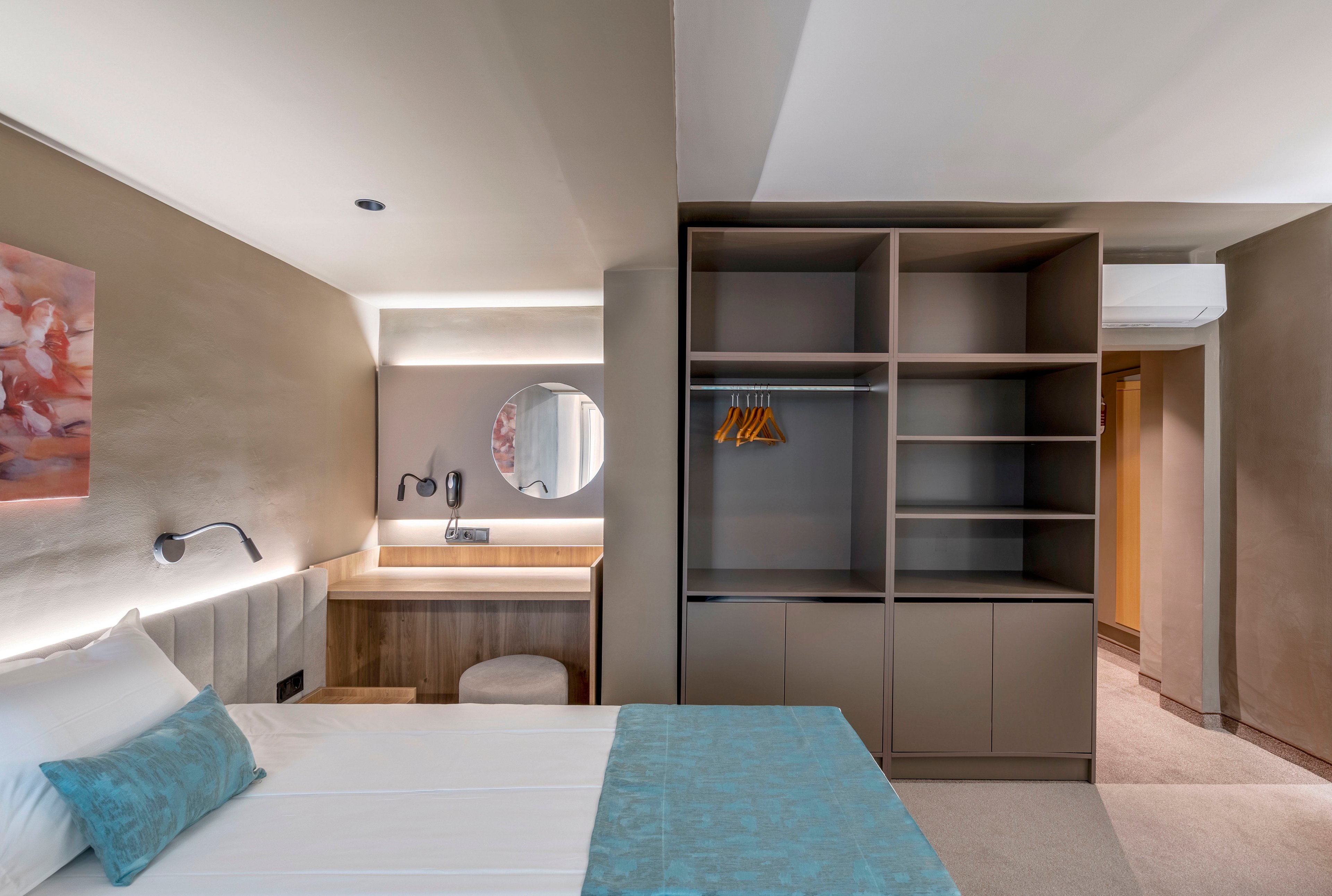 Contemporary hotel room interior featuring a neatly made bed with white linens and a turquoise runner. An open wardrobe with shelves and hanging space stands beside a compact wooden desk and round mirror. Soft wall lighting, neutral beige tones, and minimal decor create a calm, inviting atmosphere. The space appears clean, organized, and designed for comfortable short stays.