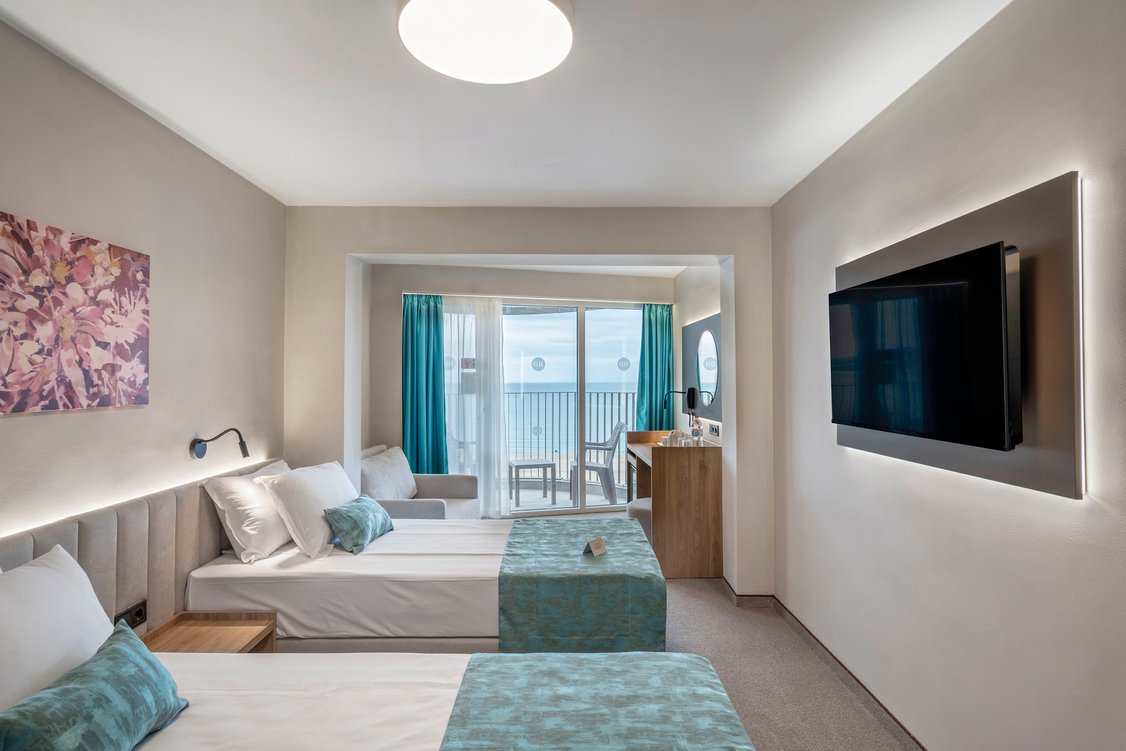 Bright contemporary hotel room featuring two neatly made twin beds with teal accents. A large flat-screen television is mounted on the wall opposite the beds, framed by subtle ambient lighting. Floor-to-ceiling glass doors open to a balcony with outdoor seating and a view of the sea. Soft neutral walls, a floral wall art piece, and turquoise curtains create a calm, coastal atmosphere.