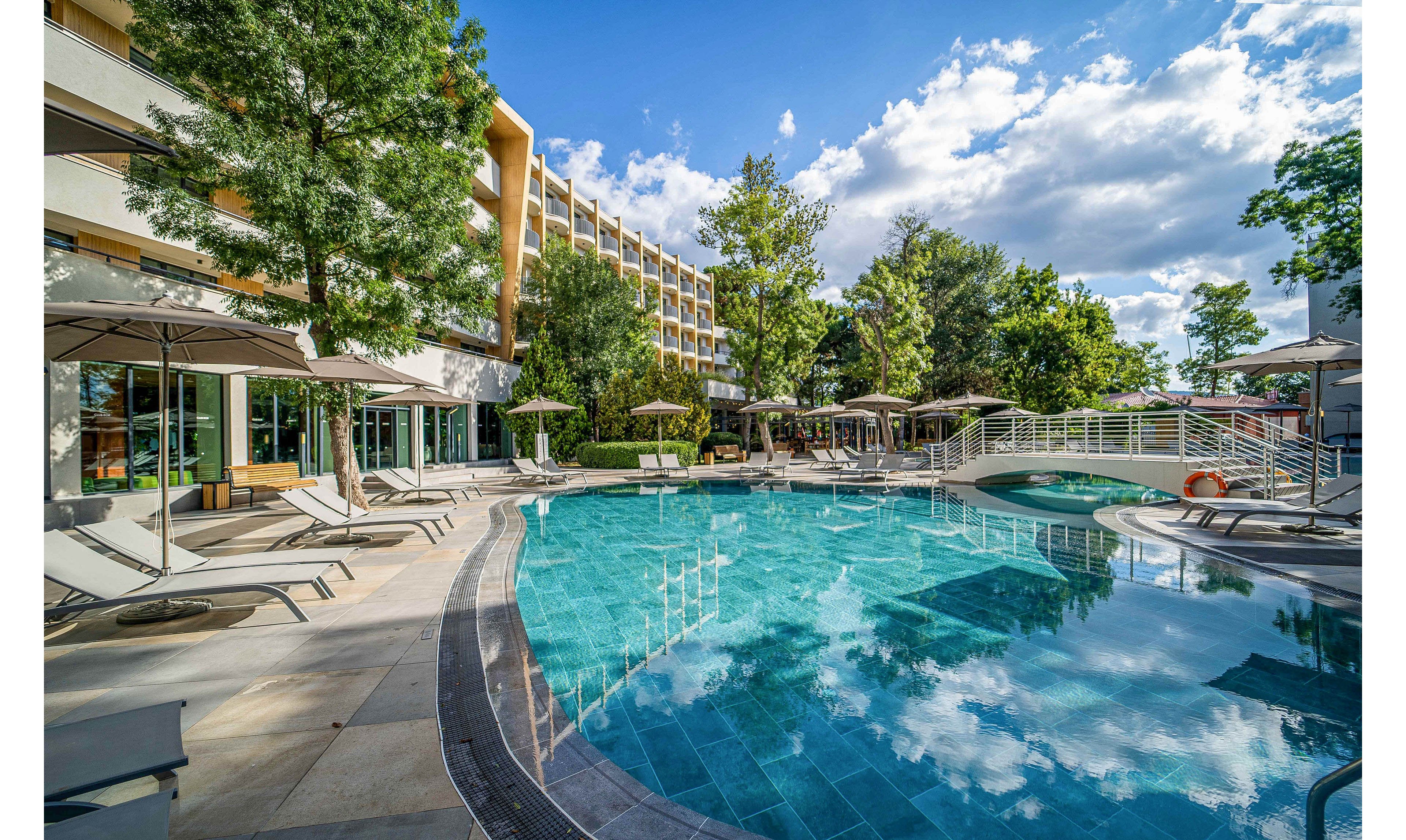A large curved swimming pool sits at the center of a modern hotel courtyard. Sun loungers and umbrellas line the poolside, creating a relaxing resort atmosphere. A small bridge crosses part of the pool, and tall trees add greenery around the area. The multi-story hotel building and bright blue sky with scattered clouds complete the sunny vacation setting.