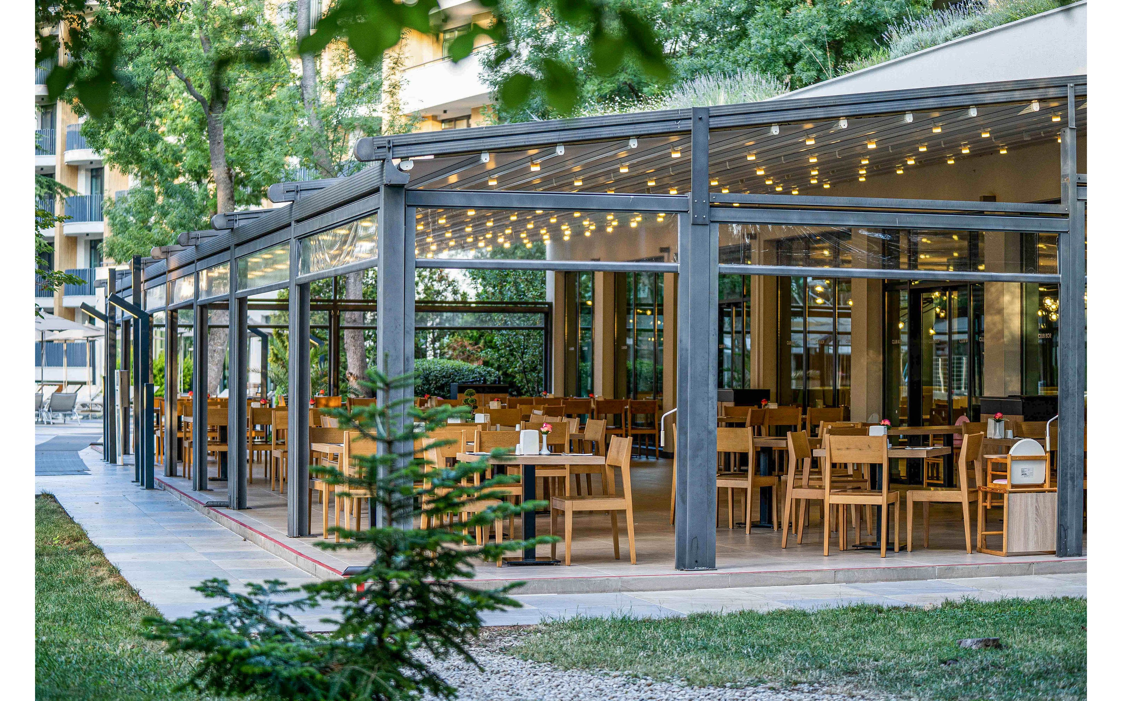 An open-air restaurant terrace features rows of wooden tables and chairs under a modern pergola structure. Warm string lights hang from the ceiling, creating an inviting ambiance in the dining area. The space is surrounded by greenery, including trees and small shrubs, with a paved walkway leading past the seating. Large glass panels enclose parts of the terrace, blending indoor comfort with an outdoor garden setting.