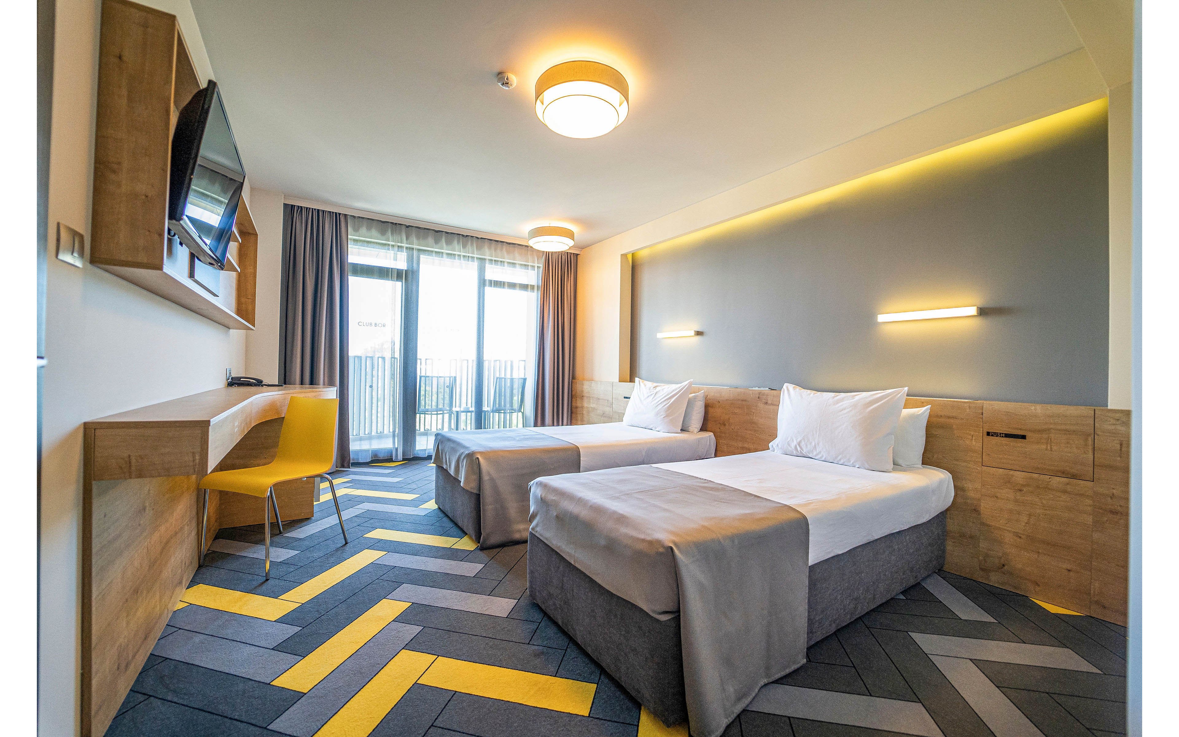 Contemporary hotel room featuring two neatly made single beds with white linens and grey bases. The space includes a wall-mounted television, wooden desk with a yellow chair, and built-in bedside storage. Large floor-to-ceiling windows with sheer and blackout curtains open to a balcony, allowing natural light to fill the room. Geometric patterned flooring in grey tones with yellow accents and minimalist wall lighting create a clean, modern atmosphere.