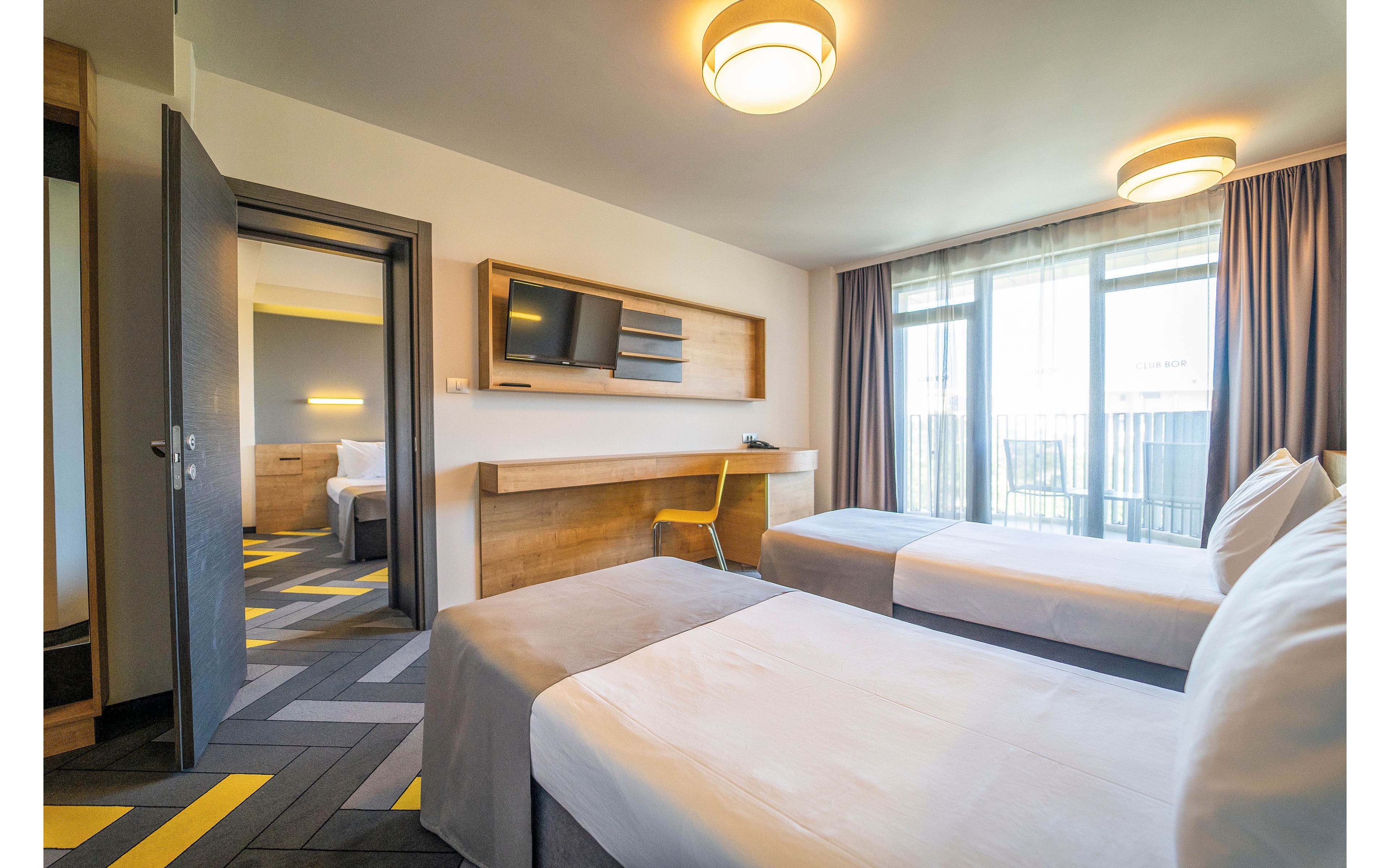 Contemporary hotel room featuring two neatly made twin beds and large windows with sheer curtains. A wall-mounted television, long wooden desk, and yellow chair create a functional workspace. The open door reveals a connecting bedroom with similar decor and geometric patterned flooring. Soft lighting and neutral tones give the space a clean, comfortable, and inviting atmosphere.