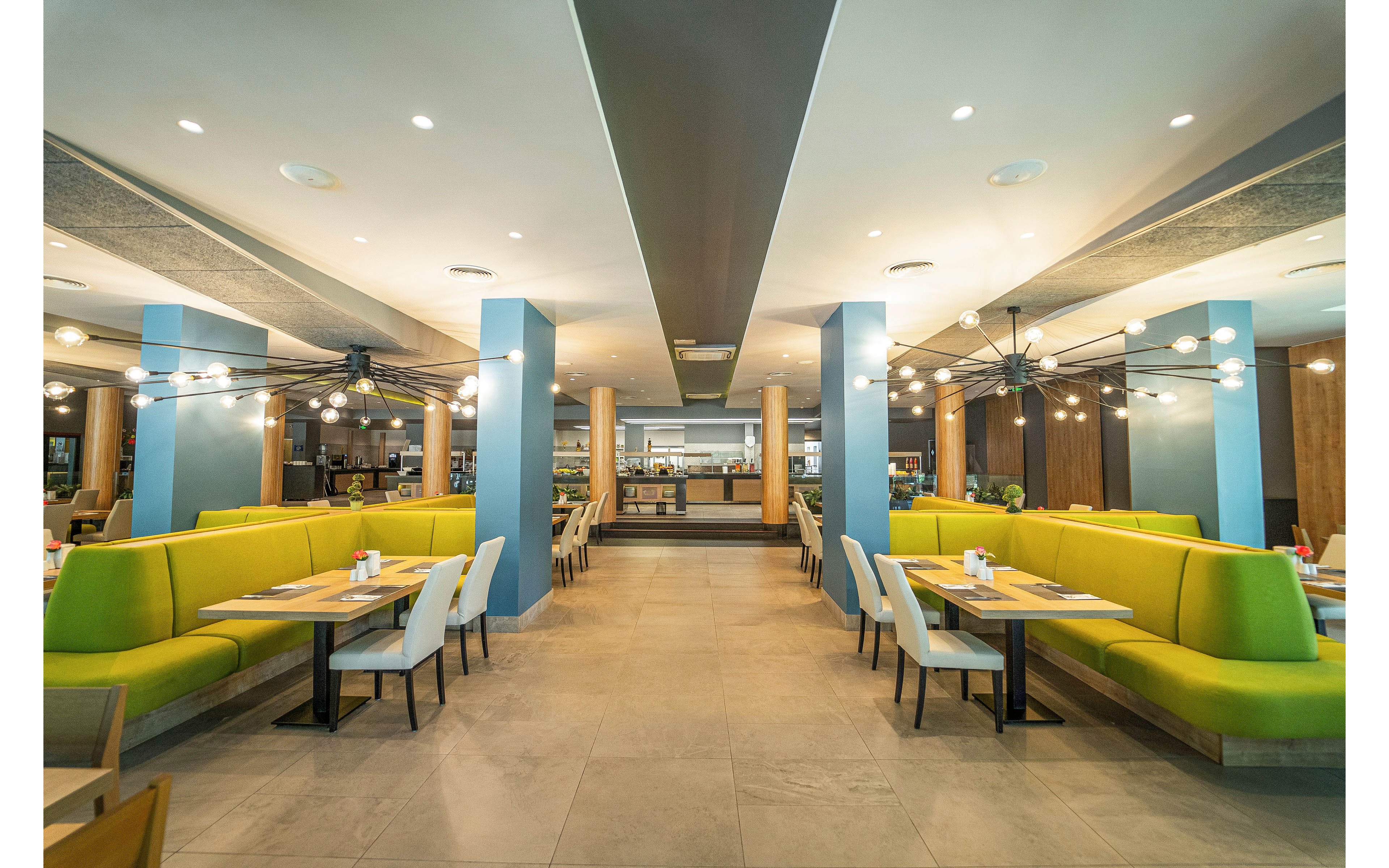 Wide view of a contemporary restaurant interior featuring bright green booth seating and light-colored chairs. The space is empty of people, emphasizing the clean, organized dining area. Blue columns and large ceiling light fixtures create a modern, symmetrical look. Warm wood accents and neutral floor tiles contribute to a welcoming, casual dining atmosphere.