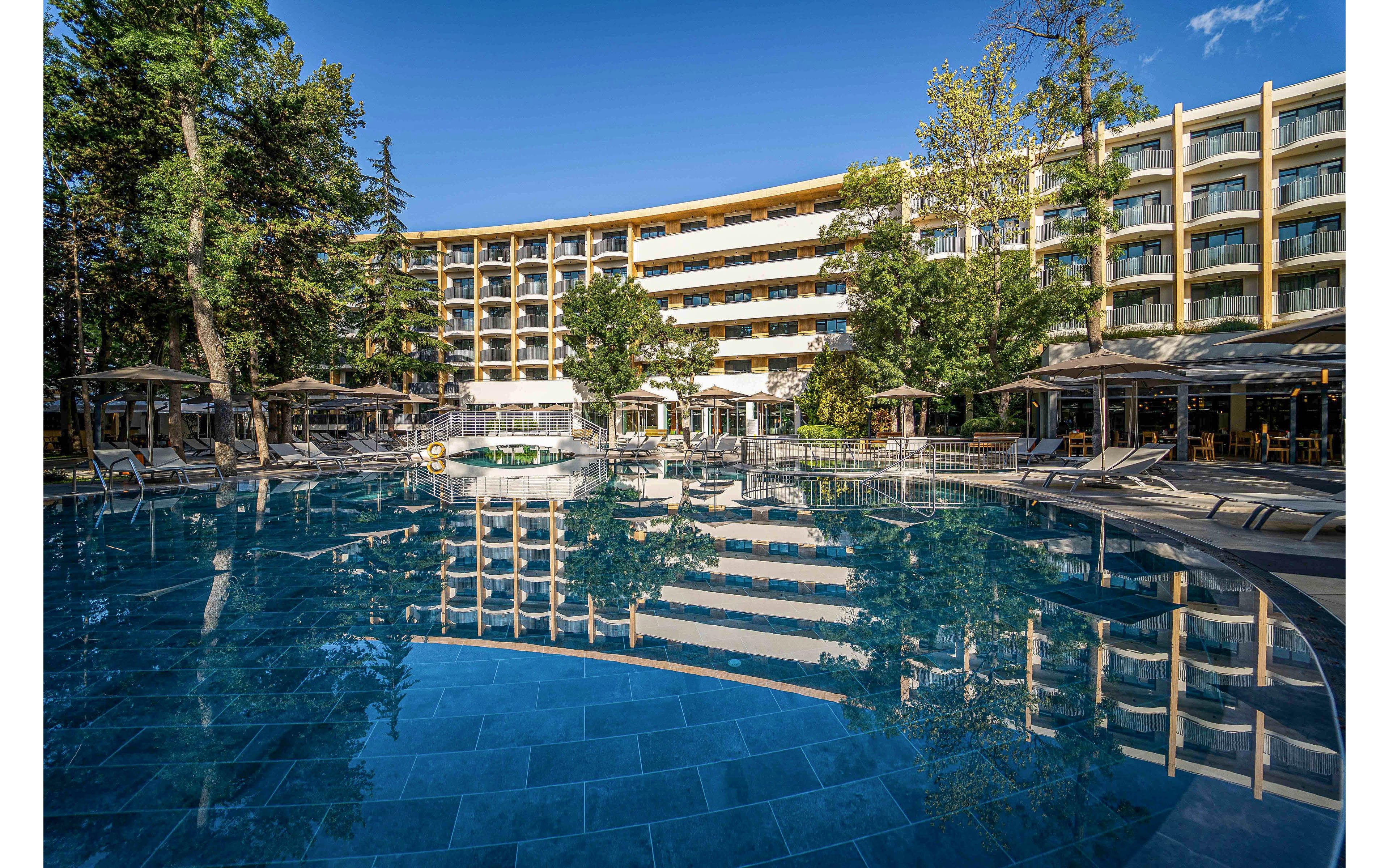 A large curved resort hotel overlooks a clear blue outdoor swimming pool. Sun loungers and umbrellas are arranged neatly around the poolside, suggesting a relaxing vacation setting. Tall trees and greenery surround the property, adding a natural backdrop to the modern architecture. The bright daylight and clear sky create a warm, inviting atmosphere for guests.