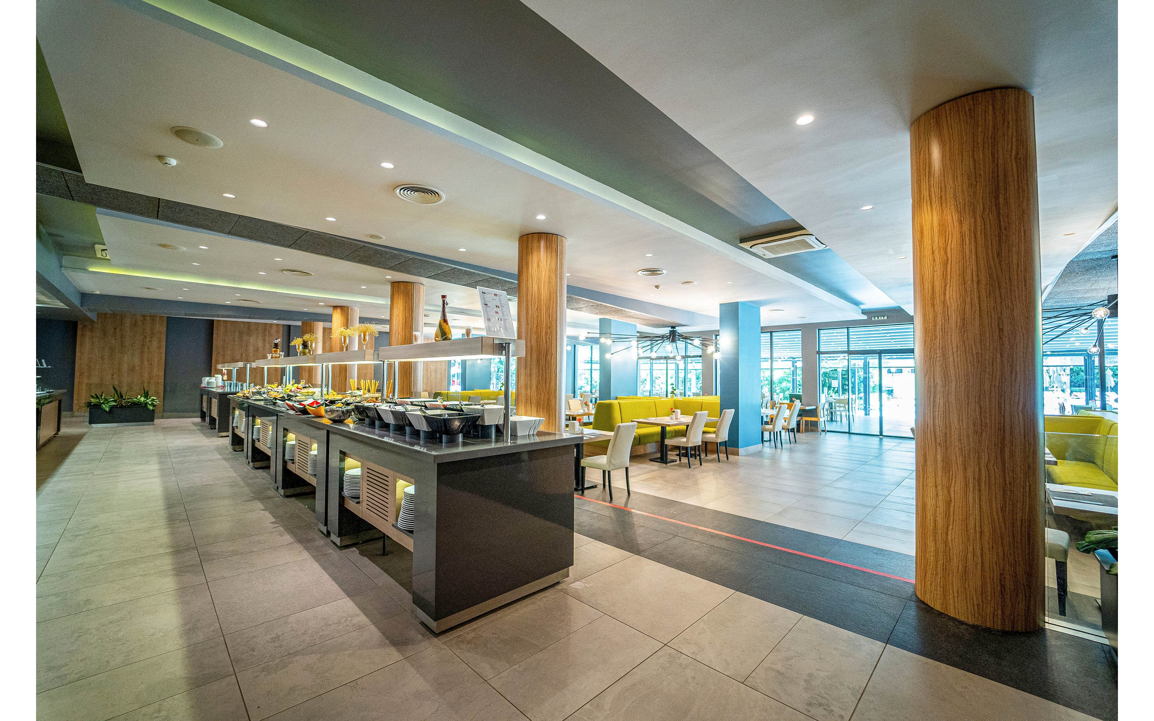 Spacious self-service buffet area in a contemporary restaurant or hotel dining room. Multiple food stations are arranged in a row with warm lighting and sleek counters. Wooden columns, tiled floors, and large windows create an open, airy atmosphere. Green and yellow upholstered chairs surround tables near the windows, suggesting a comfortable place for guests to dine.