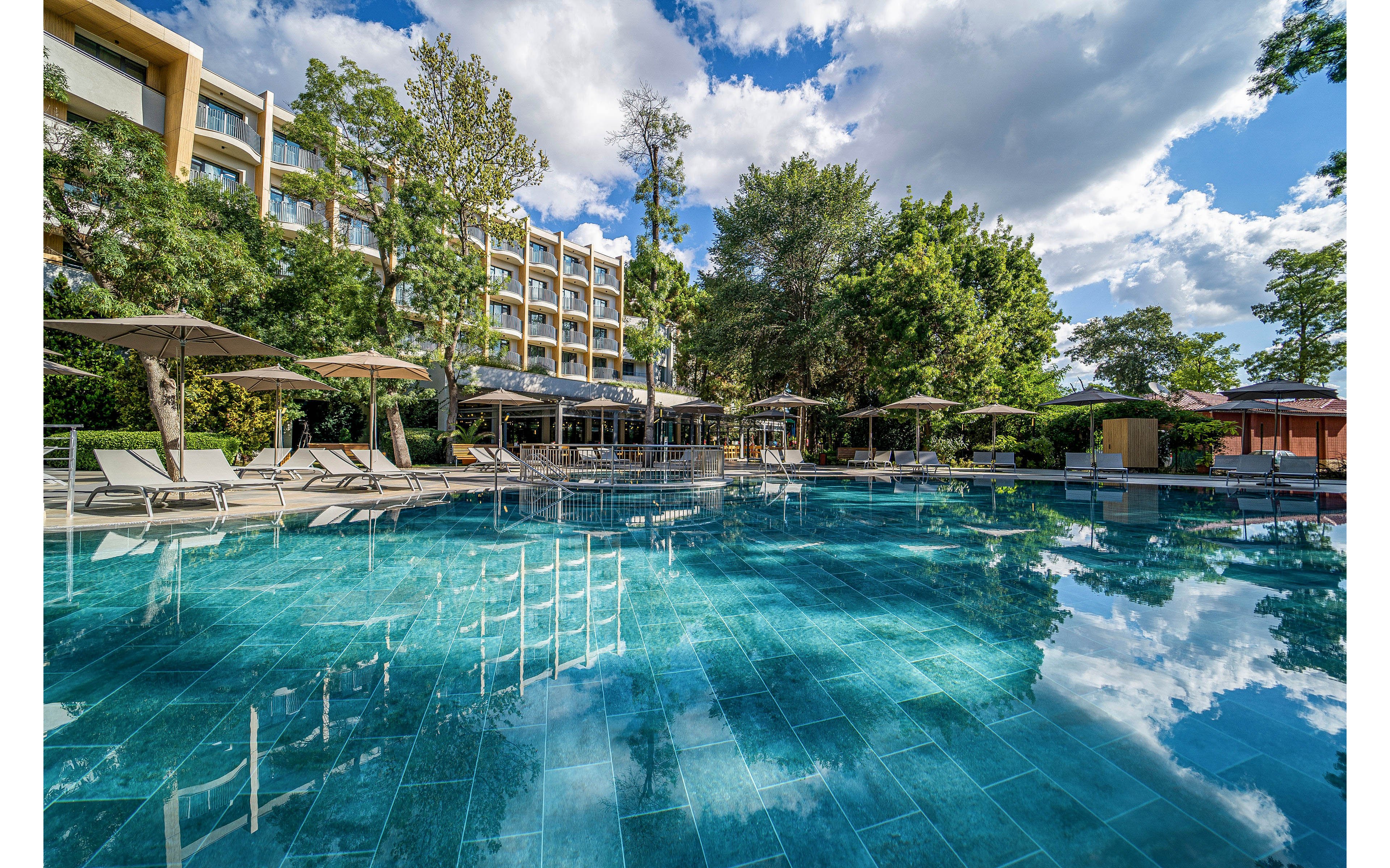 A large, clear blue swimming pool sits in front of a multi-story hotel surrounded by lush green trees. Sun loungers and umbrellas line the poolside, creating a relaxing resort atmosphere. Reflections of the sky, clouds, and trees shimmer on the water’s surface. The scene suggests a tranquil vacation setting on a bright, partly cloudy day.