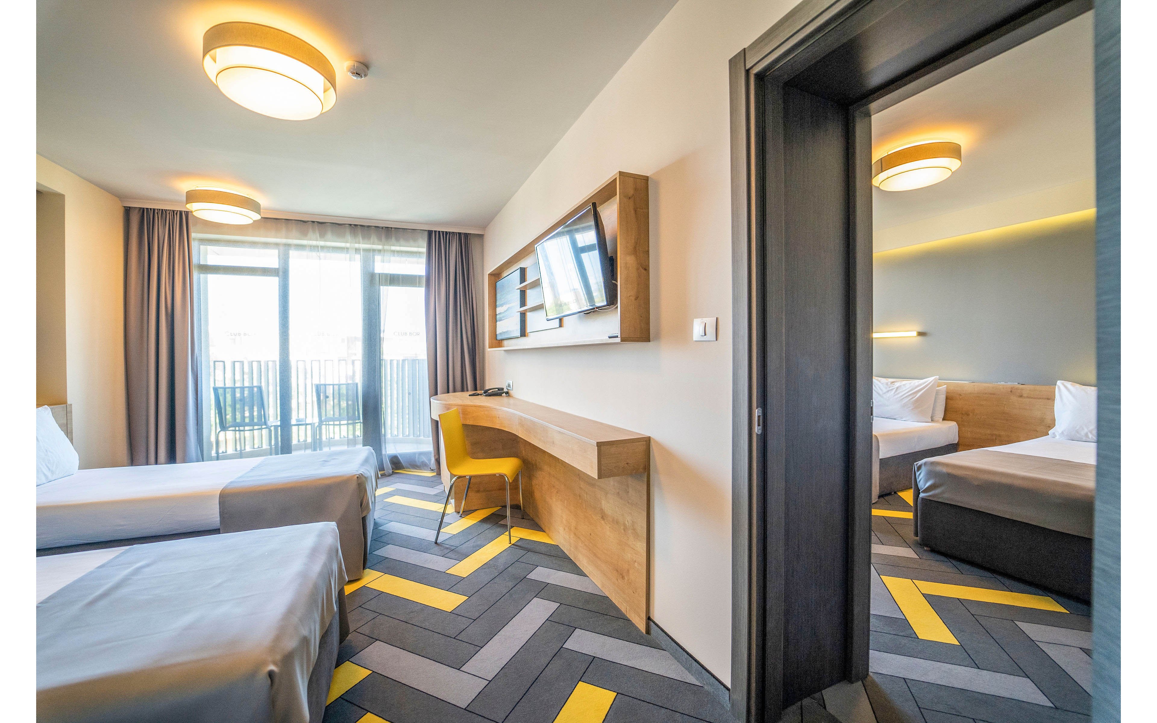 Contemporary hotel room interior featuring two neatly made twin beds and a sleek wooden desk with a yellow chair. Large windows with curtains allow natural light to brighten the space and offer a view outside. A wall-mounted television and built-in shelving add functional comfort to the room. An adjoining bedroom with a double bed is visible through an open doorway, emphasizing a spacious, connected suite layout.