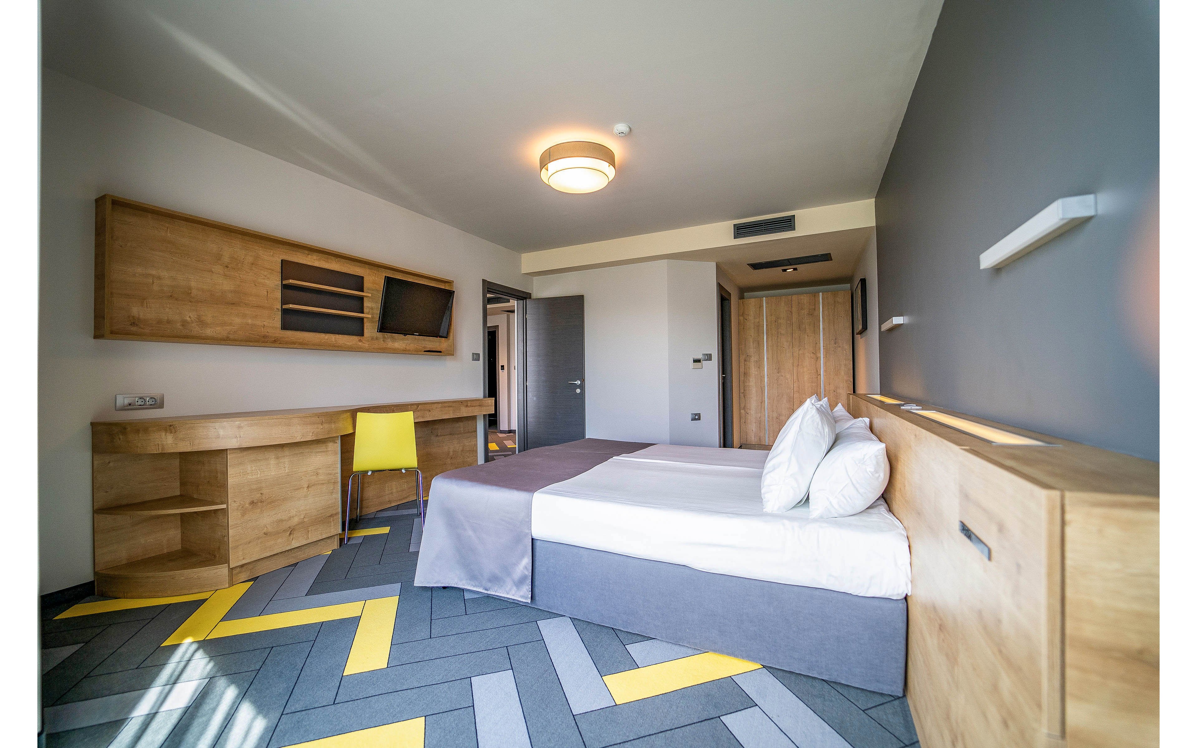 Contemporary hotel room interior featuring a neatly made double bed with white pillows and a gray throw. The space includes built-in wooden furniture, a long desk, wall shelving, and a mounted television. A bright yellow chair adds a pop of color against the neutral gray and beige walls. The patterned floor with gray and yellow chevron accents and a ceiling light fixture complete the modern, minimalist design.