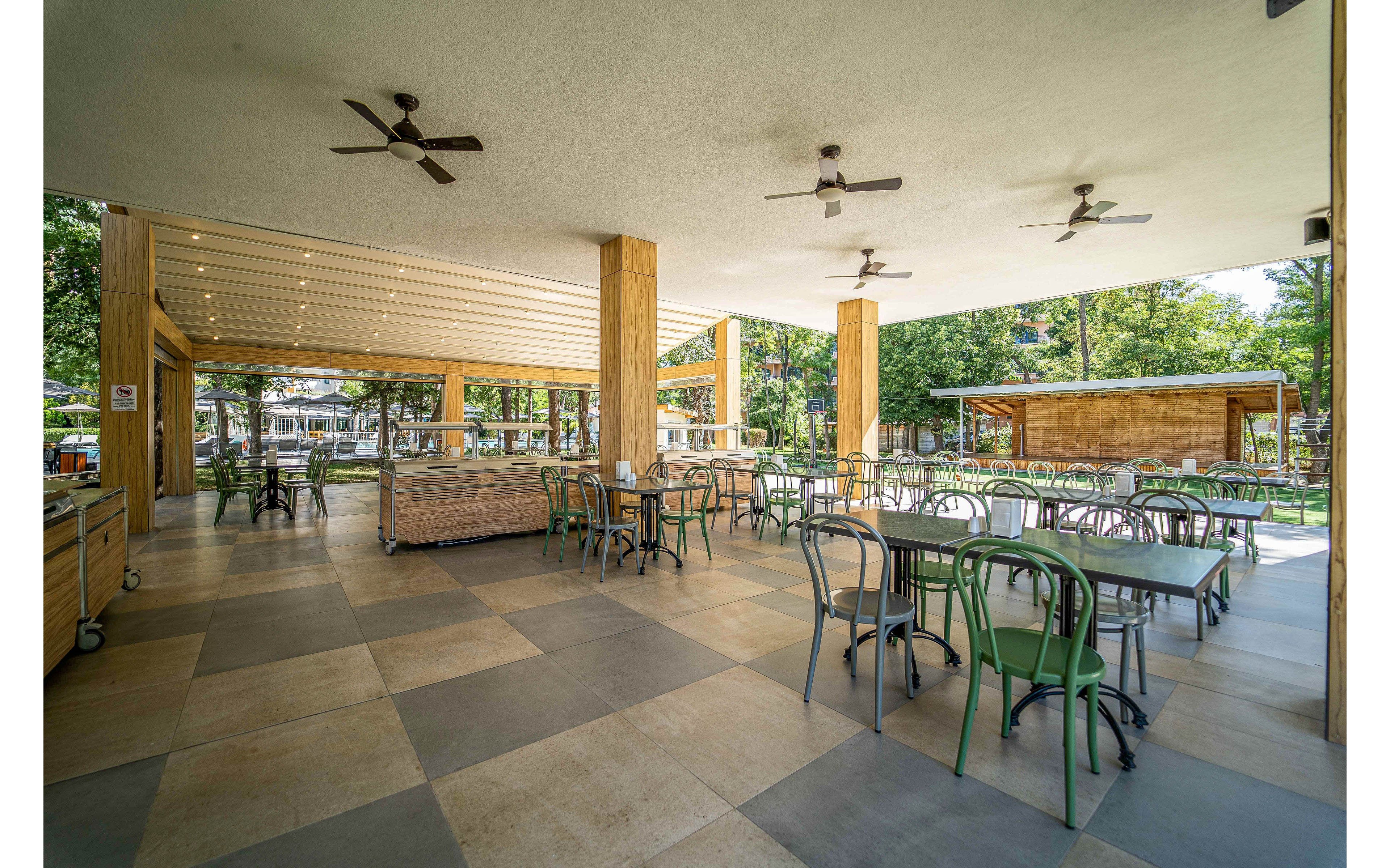 Spacious open-air dining area features rows of empty tables and metal chairs under a covered patio. Wooden columns and serving counters frame the space, with ceiling fans and recessed lighting overhead. Large openings reveal a green garden setting with trees and additional structures in the background. The scene appears calm and ready for guests, suggesting a restaurant, café, or resort dining space.