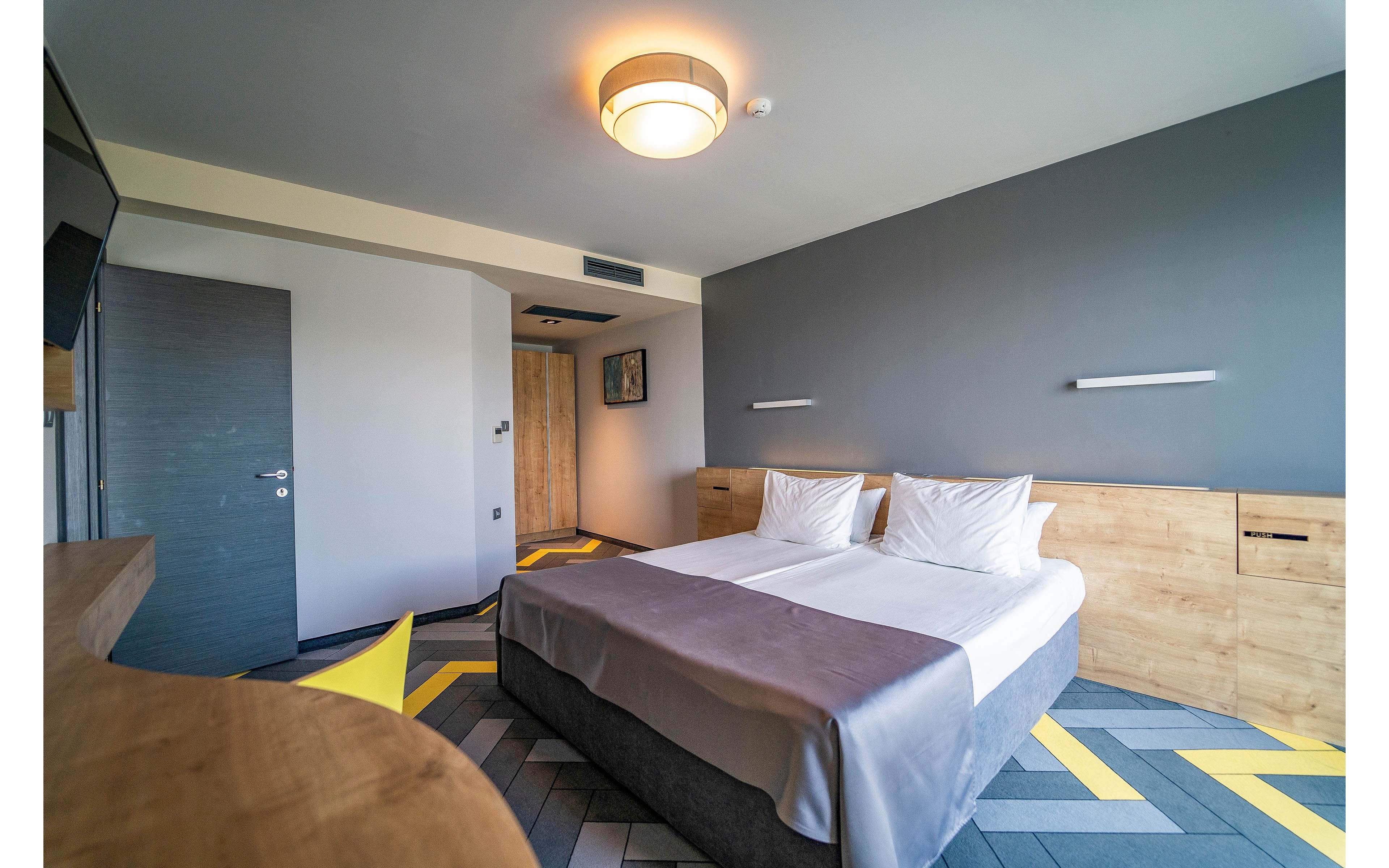 Contemporary hotel room interior featuring two neatly made twin beds with white linens and gray bed runners. The space has minimalist wooden furniture, including a headboard, side cabinets, and a desk. A geometric patterned floor in gray and yellow adds visual interest to the neutral gray walls. Soft ceiling lighting and wall-mounted reading lights create a calm, inviting atmosphere.