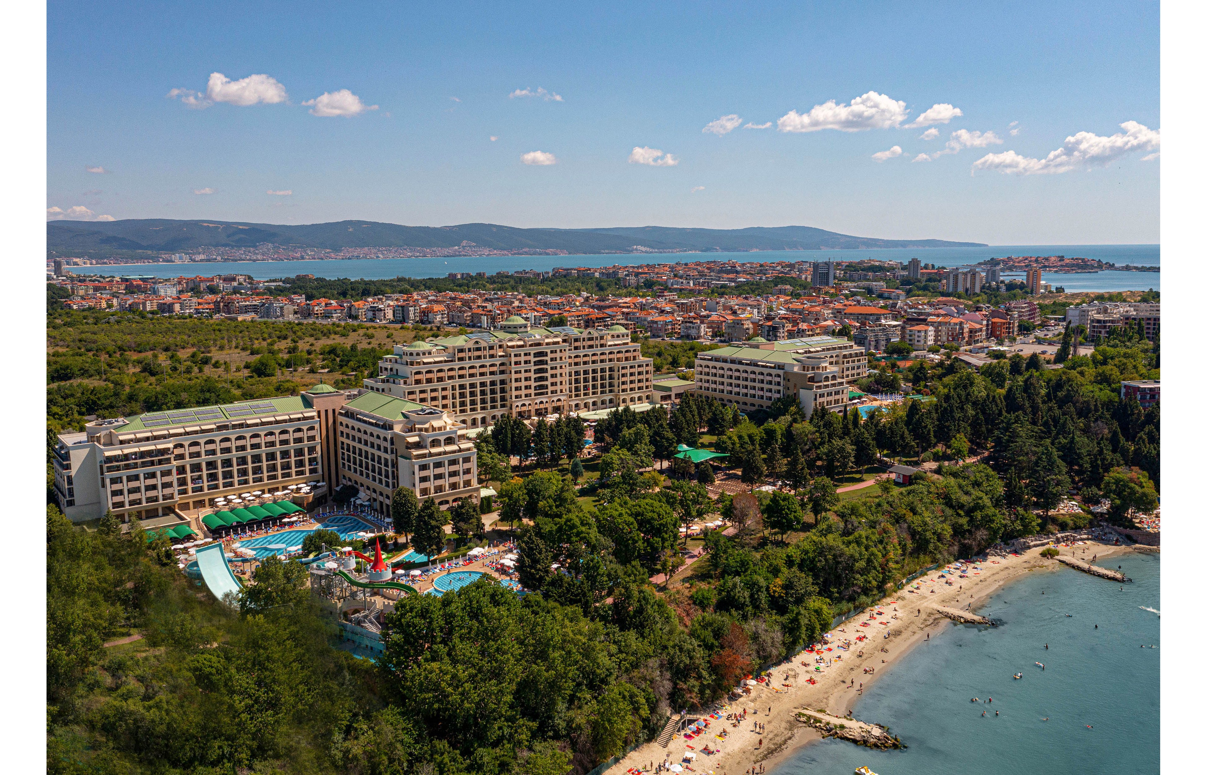 A large coastal resort complex with multiple hotel buildings and pools is seen from above along a sandy beach. The shoreline is dotted with sunbathers and umbrellas, while lush green trees separate the resort from the water. In the background, a sprawling city extends toward distant hills and a calm bay. The scene is bright and sunny, suggesting a warm summer vacation atmosphere.