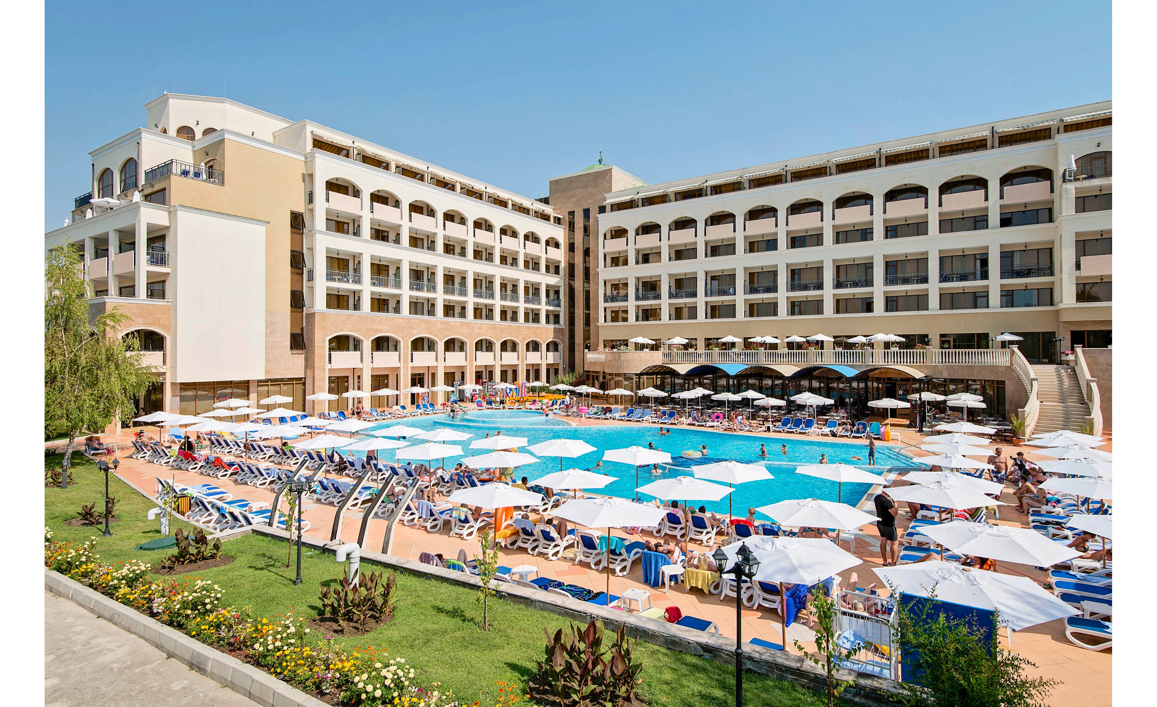 A large multi-story resort hotel surrounds a central outdoor swimming pool filled with guests. Numerous white and blue sun loungers and umbrellas line the pool deck, creating a busy vacation atmosphere. The building features balconies on each floor and arched ground-level openings facing the pool. In the foreground, a landscaped garden with flowers and greenery borders the pool area. The scene is brightly lit under clear blue skies, suggesting a warm, sunny day.