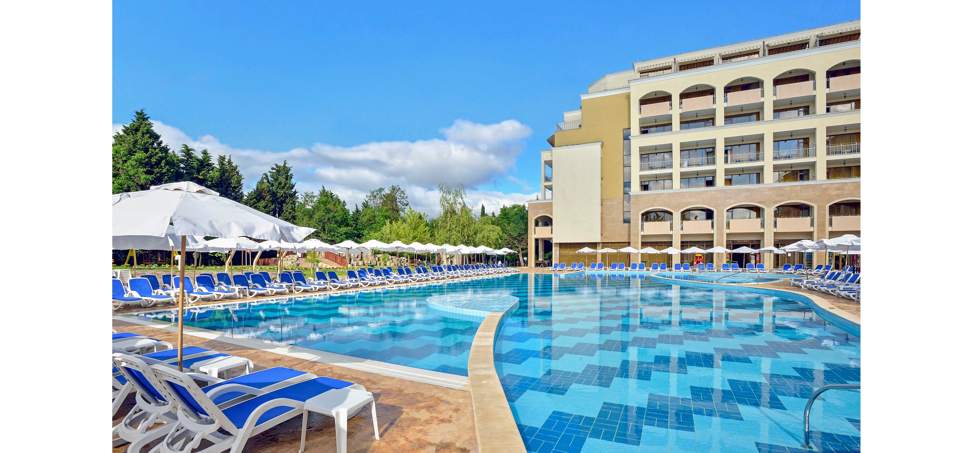 Large outdoor swimming pool reflects a multi-story resort hotel under a bright blue sky. Numerous white sun loungers with blue cushions line the poolside, shaded by white umbrellas. The hotel features balconies and arched windows overlooking the pool area. Green trees frame the scene, adding a natural backdrop to the leisure setting.