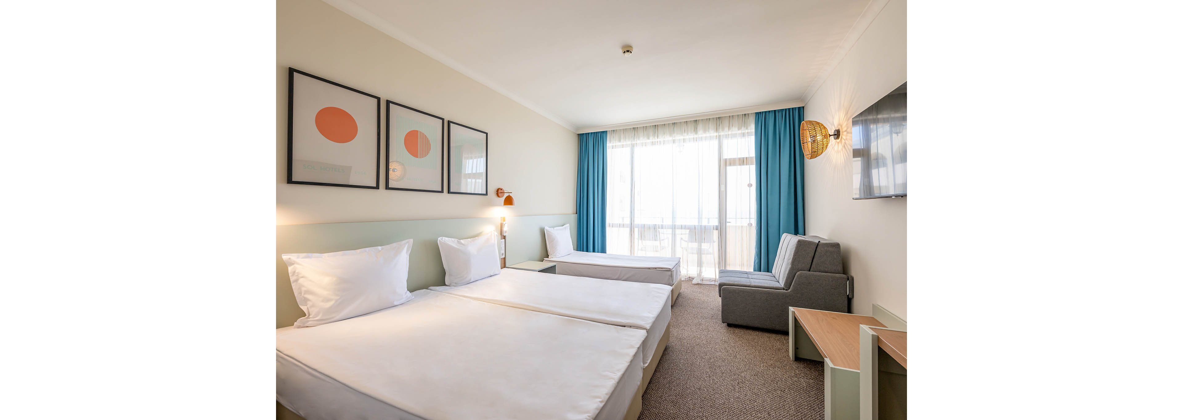 A bright, modern hotel room features two neatly made twin beds with white linens in the foreground. A third bed or sofa bed sits near the window, alongside a gray armchair and small wooden table. Large windows with sheer curtains and blue drapes let in abundant natural light, revealing a balcony outside. Minimalist wall art, warm wall sconces, and neutral tones create a clean, inviting atmosphere suitable for travelers or guests.