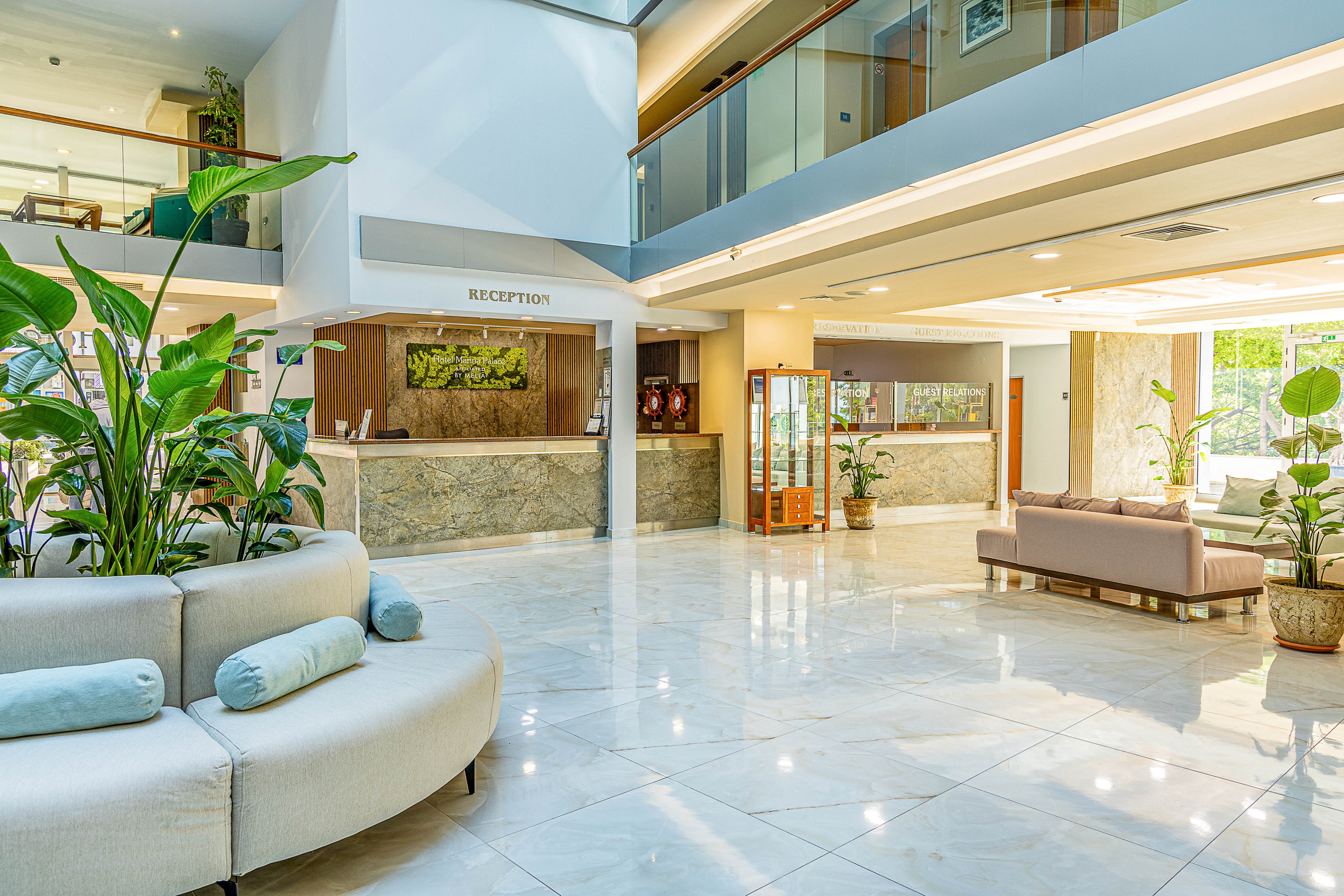 Spacious hotel lobby interior featuring a central curved sofa and lush green plants. A marble-front reception desk sits in the background beneath a sign reading RECEPTION. Large windows and glass railings on the upper floor allow abundant natural light to fill the space. Soft neutral tones, polished floors, and contemporary furniture create a clean, welcoming atmosphere.
