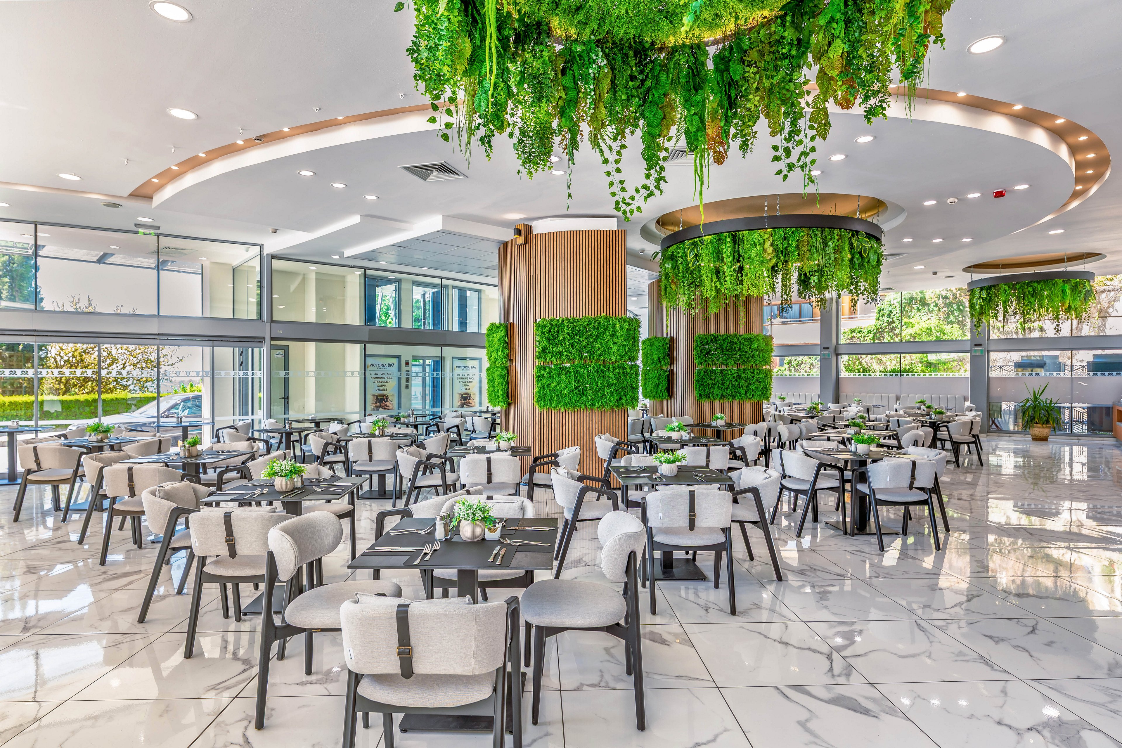 Spacious contemporary restaurant interior features numerous dining tables arranged on a glossy white marble floor. Large windows and glass doors flood the space with natural daylight and views of the outdoor greenery. Cylindrical columns and ceiling fixtures are covered with cascading green plants, creating a fresh, biophilic atmosphere. Neutral-toned chairs and minimalist table settings contribute to the clean, upscale design.