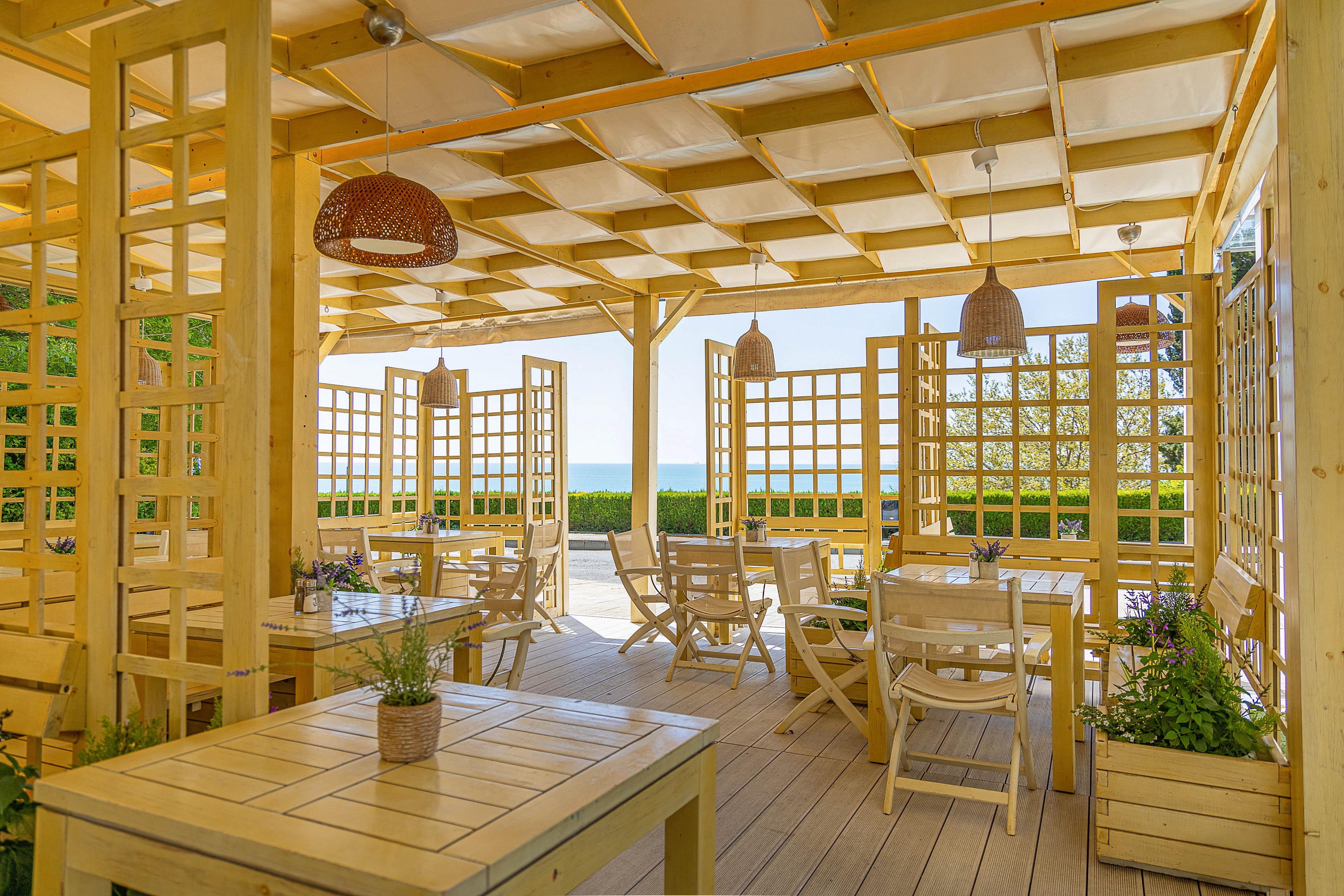 Open-air café terrace with light wooden tables and chairs sits under a structured pergola roof. Decorative lattice panels divide the seating area, allowing views of greenery and the distant sea horizon. Potted plants and woven pendant lights add natural accents and warmth to the space. Bright daylight and soft shadows create a relaxed, summery coastal atmosphere.