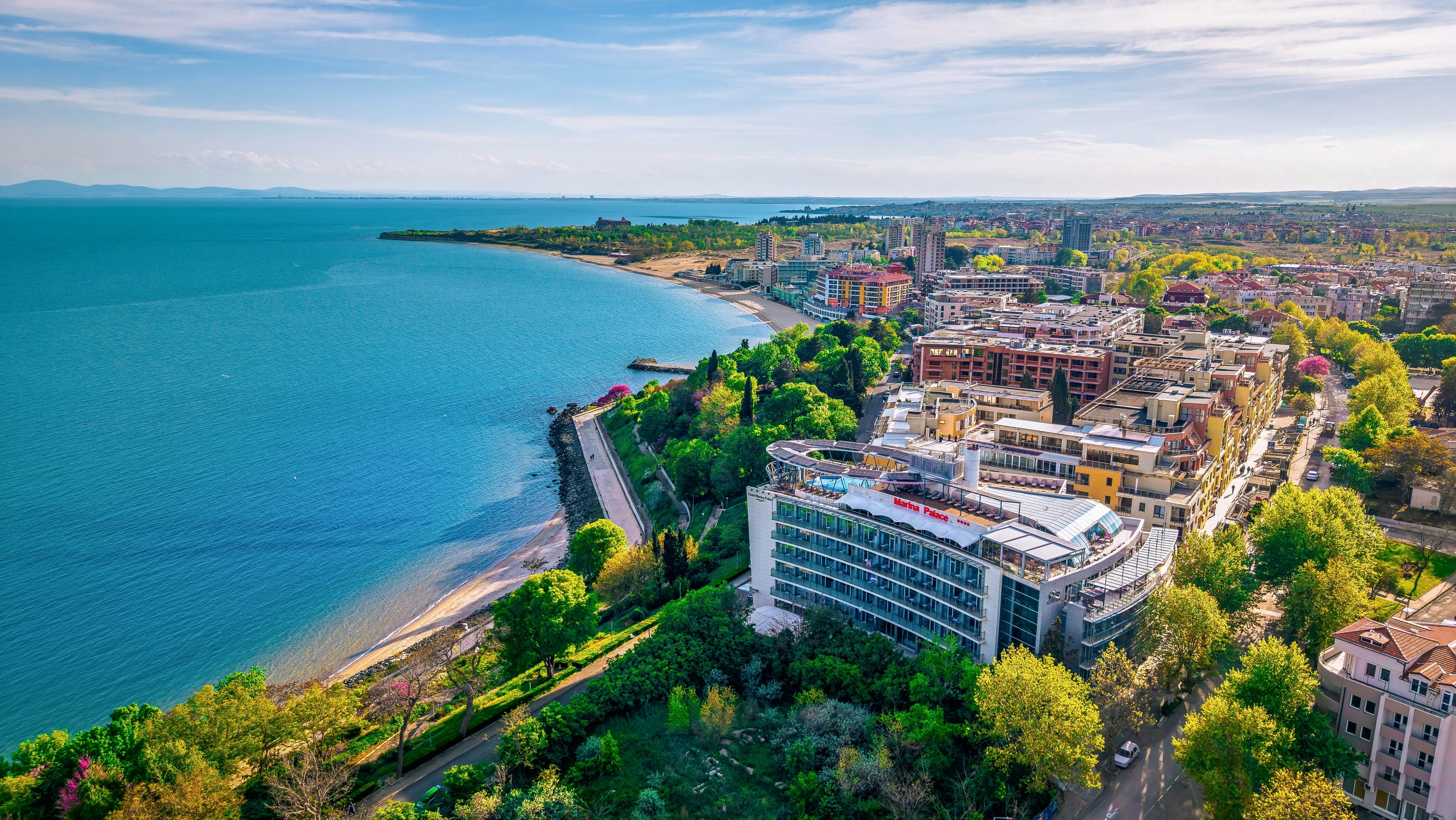 Aerial view of a coastal city with a sweeping blue bay on the left and dense urban development on the right. Modern multi-story buildings line the waterfront, surrounded by lush green trees and park areas. A curving shoreline road separates the city from the calm sea. The scene is captured on a clear day with bright, vibrant colors and a wide horizon in the distance.