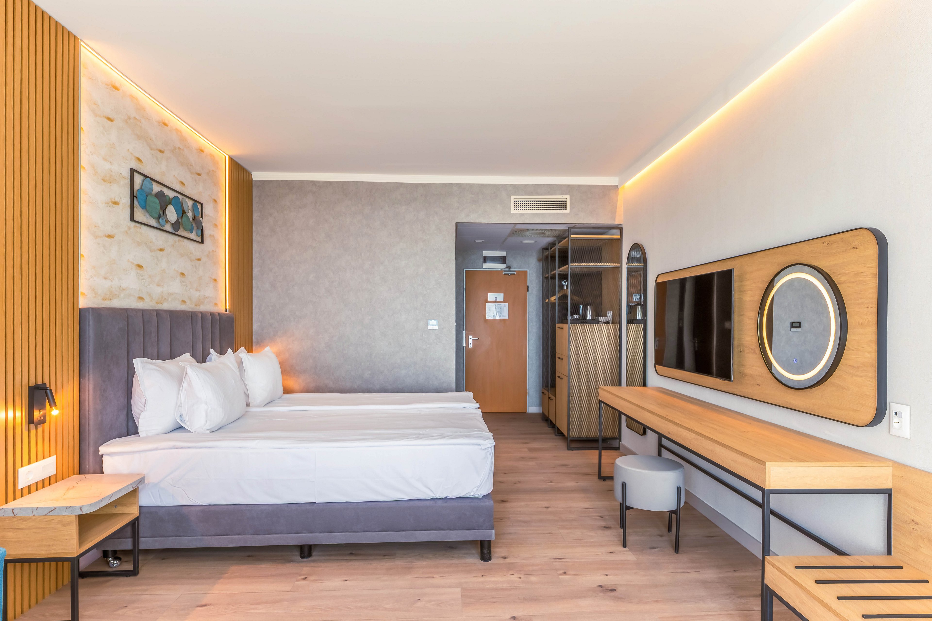 Contemporary hotel room interior featuring two neatly made double beds with white linens. The space includes a wall-mounted flat-screen TV, long wooden desk, and small upholstered stool. Warm accent lighting and wood paneling create a cozy yet minimalist atmosphere. A doorway, wardrobe, and small kitchenette area are visible in the background, emphasizing comfort and functionality.