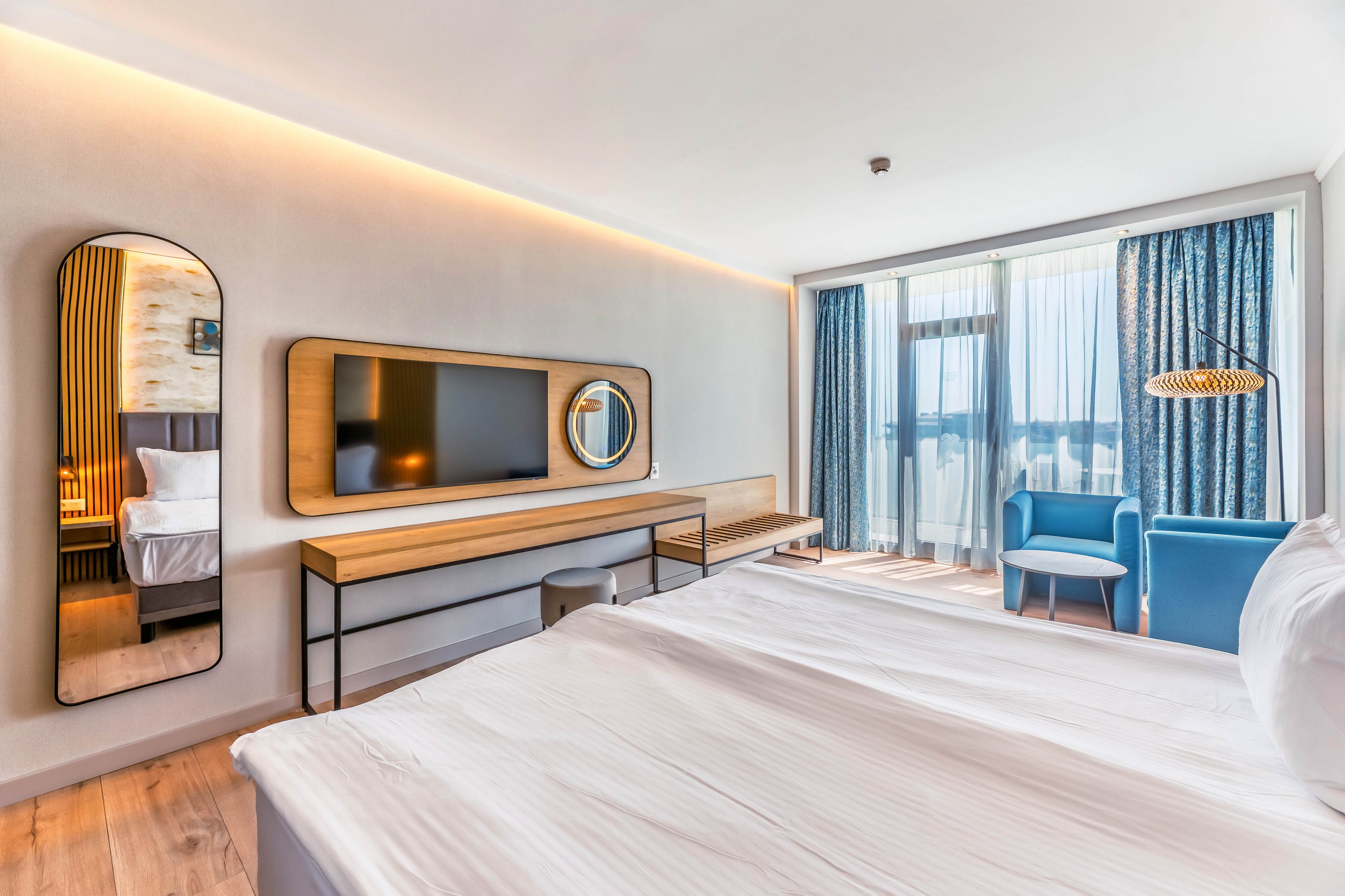 A contemporary hotel room features a neatly made bed in the foreground facing a wall-mounted flat-screen TV. Sleek wooden furniture and two decorative mirrors add a stylish touch to the minimalist interior. Floor-to-ceiling windows with sheer and dark curtains open onto a balcony with an outdoor view. A blue armchair and small table create a cozy seating area near the natural light.
