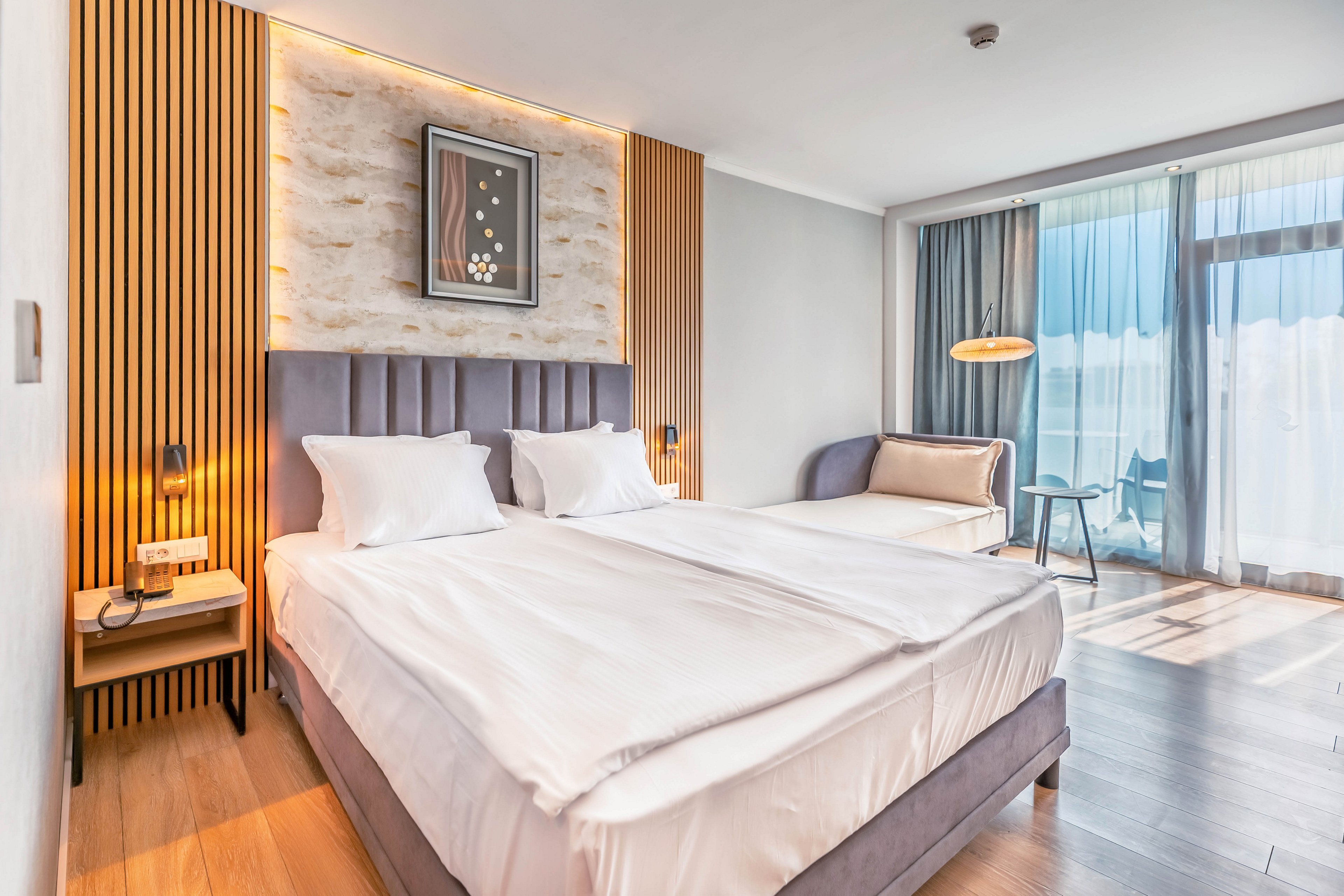 Contemporary hotel room featuring a large neatly made double bed with white linens. The space includes a small bedside table, wall-mounted lamps, and a cushioned sofa near the window. Floor-to-ceiling glass doors open to a balcony with outdoor seating and a view. Warm wood accents and soft neutral tones create a clean, inviting atmosphere suitable for travel or vacation stays.