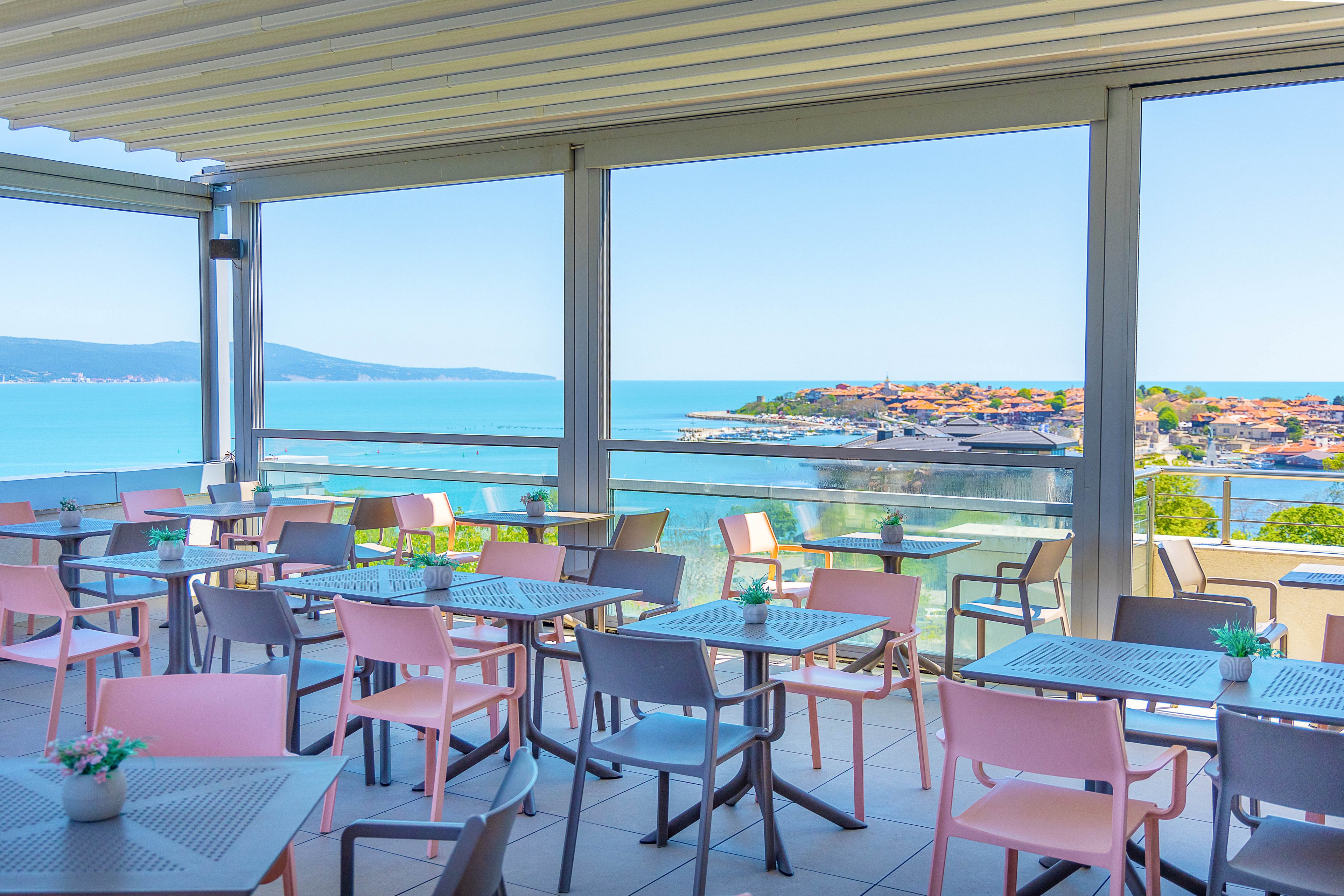 An open-air terrace cafe features colorful pastel chairs and tables arranged under a modern pergola. Large windows frame a panoramic view of a bright blue sea and a picturesque coastal town on a small peninsula. The scene is sunlit and tranquil, with clear skies and calm water creating a relaxing atmosphere. Small potted plants on the tables add a touch of greenery to the contemporary setting.