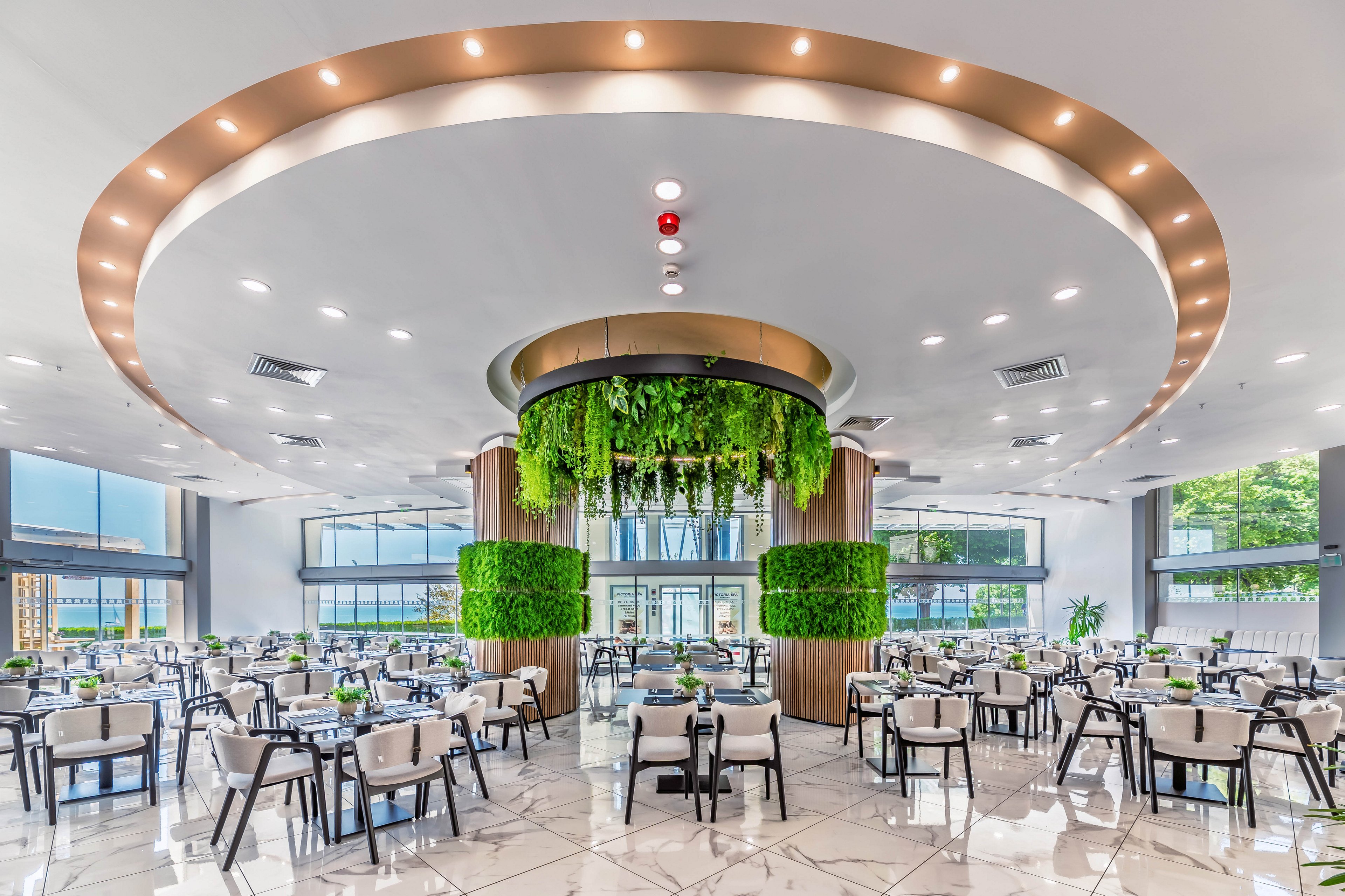 A large contemporary restaurant interior features rows of neatly arranged tables and chairs beneath a dramatic circular ceiling design. Hanging plants and vertical green walls create a lush focal point in the center of the space. Floor-to-ceiling windows bring in natural light and views of the surrounding cityscape. The bright, polished flooring and neutral furnishings contribute to a clean, upscale dining atmosphere.
