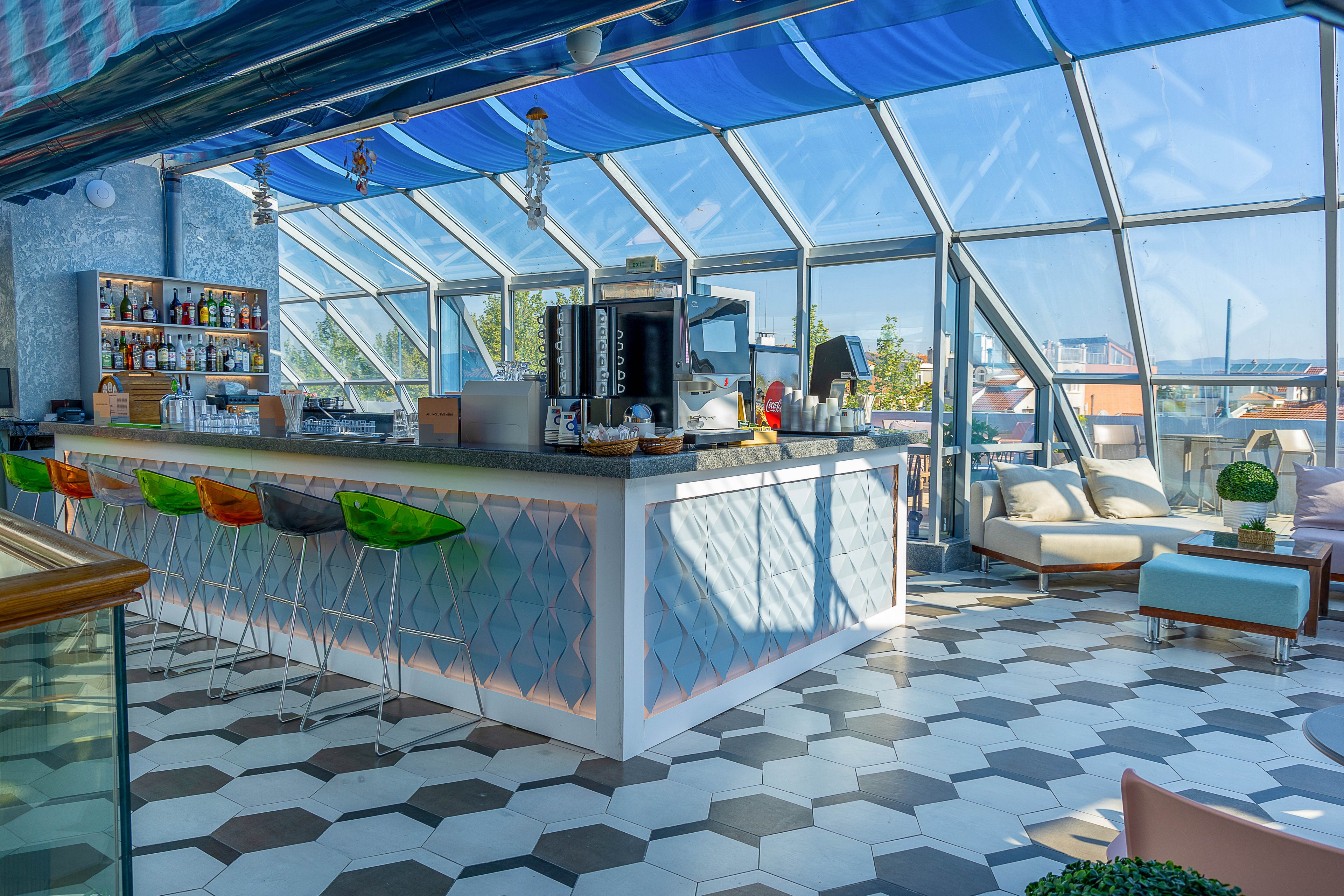 A contemporary rooftop bar features a long central counter with colorful bar stools and geometric tile patterns. Large slanted glass windows surround the space, flooding it with natural daylight and views of nearby rooftops. The bar shelves are stocked with various bottles, and a coffee machine sits on the counter. Comfortable lounge seating and potted plants create a relaxed, stylish atmosphere under a bright blue canopy.