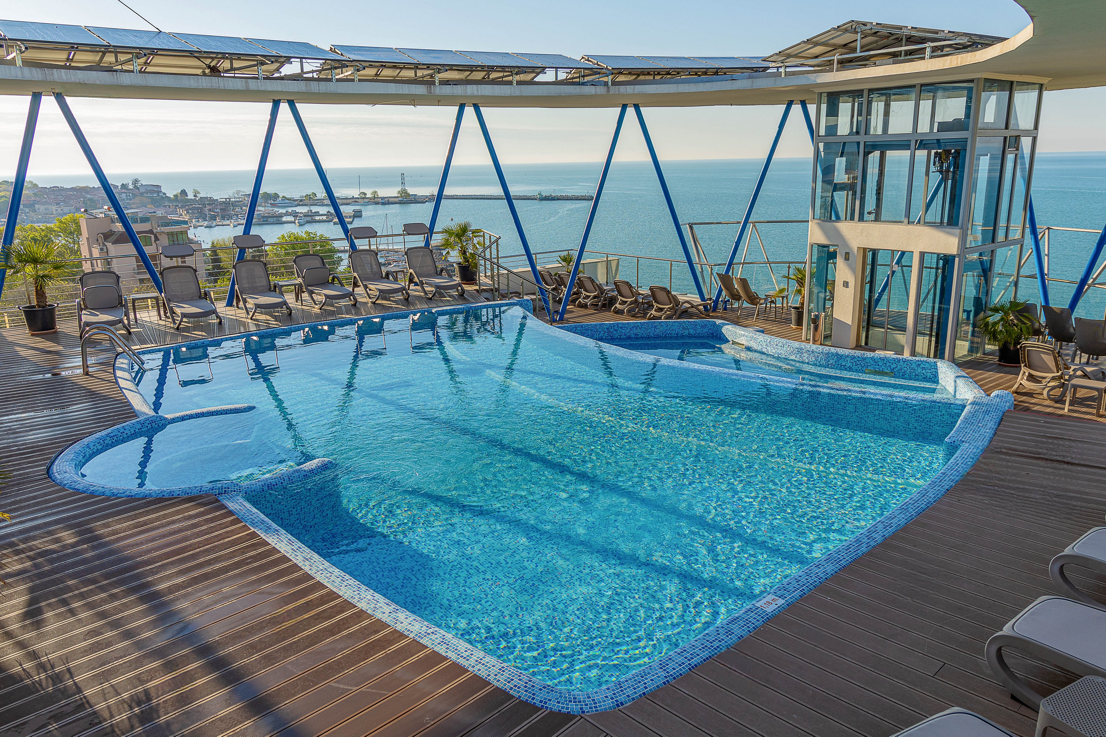 A modern rooftop infinity pool glistens under clear daylight, framed by a wooden deck and sleek metal supports. Lounge chairs and potted plants surround the pool, suggesting a relaxing leisure area in a high-rise building. Glass railings and an elevator shaft emphasize the contemporary architectural design. In the background, a coastal cityscape and calm sea stretch toward the horizon, creating a luxurious vacation atmosphere.
