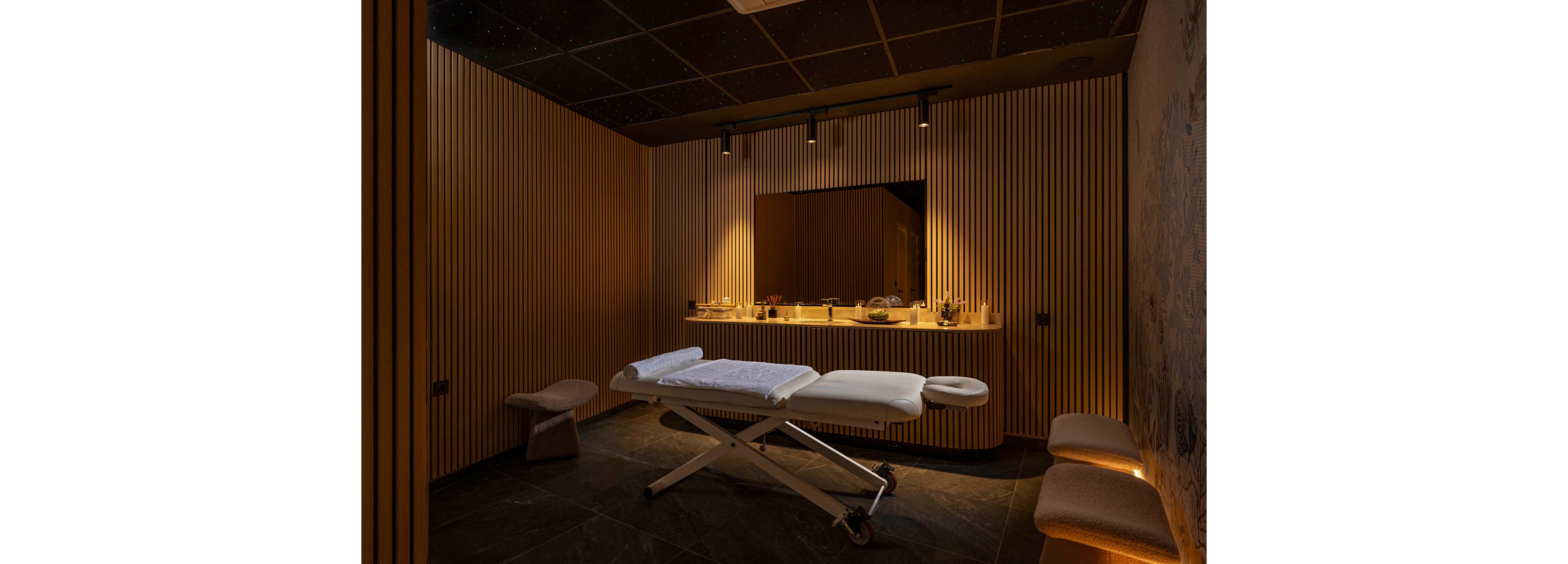 A modern spa treatment room features a central massage table covered with white linens. The space is enclosed by vertical wooden slats and a dark tiled floor, creating a cozy and intimate atmosphere. A backlit mirror and countertop with various bottles and accessories add functional elegance to the room. Soft, indirect lighting from ceiling spotlights enhances the warm, relaxing ambiance.
